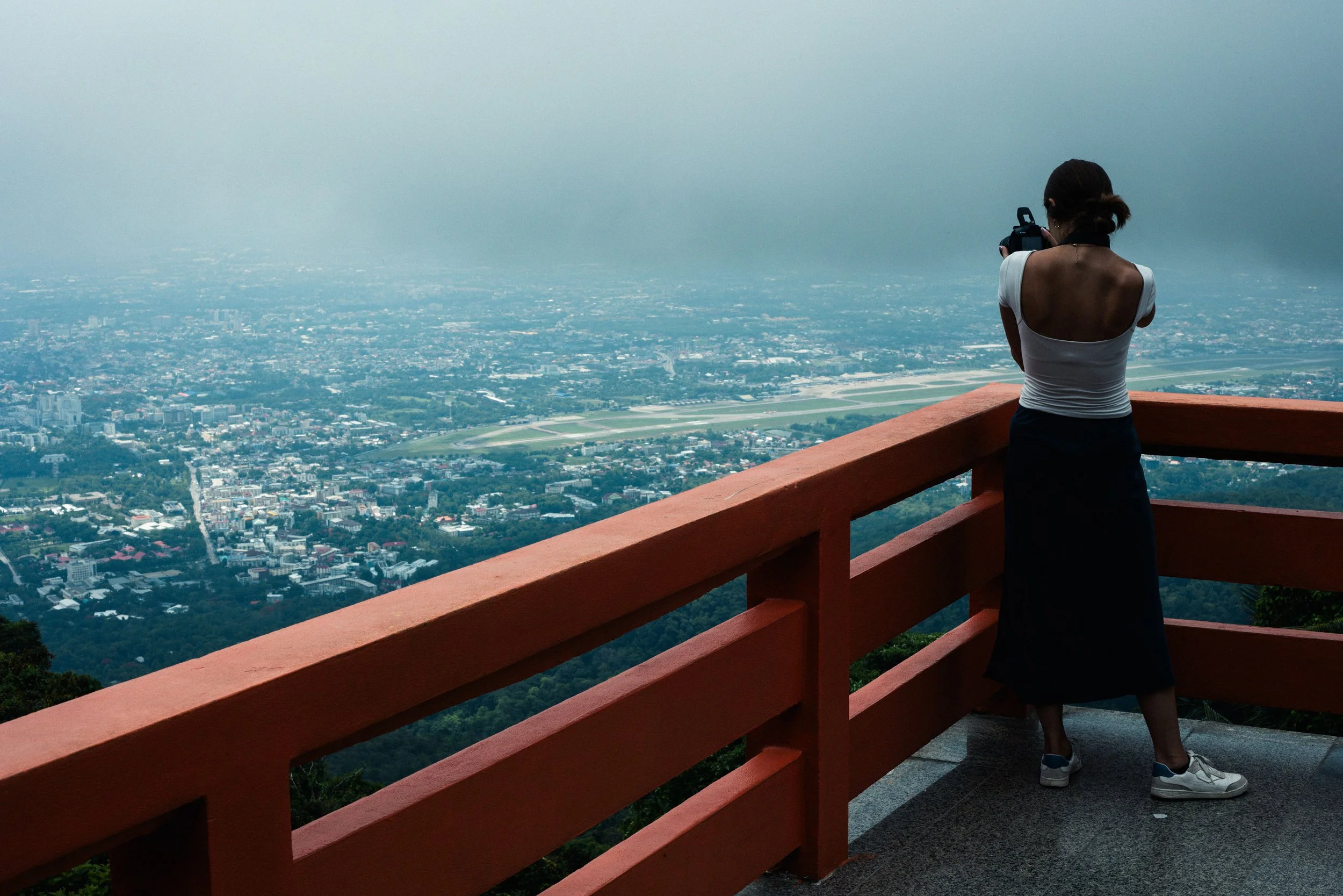 Woman taking a photo with her camera from a viewing platform overlooking a cityscape and an airport with runways, with forested hills in the foreground.