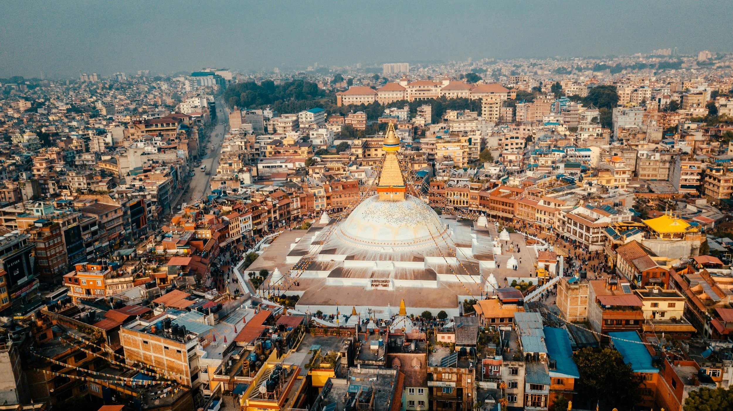 Aerial view of a city with a large white stupa at the center, surrounded by densely packed buildings and streets, with mountains in the background.