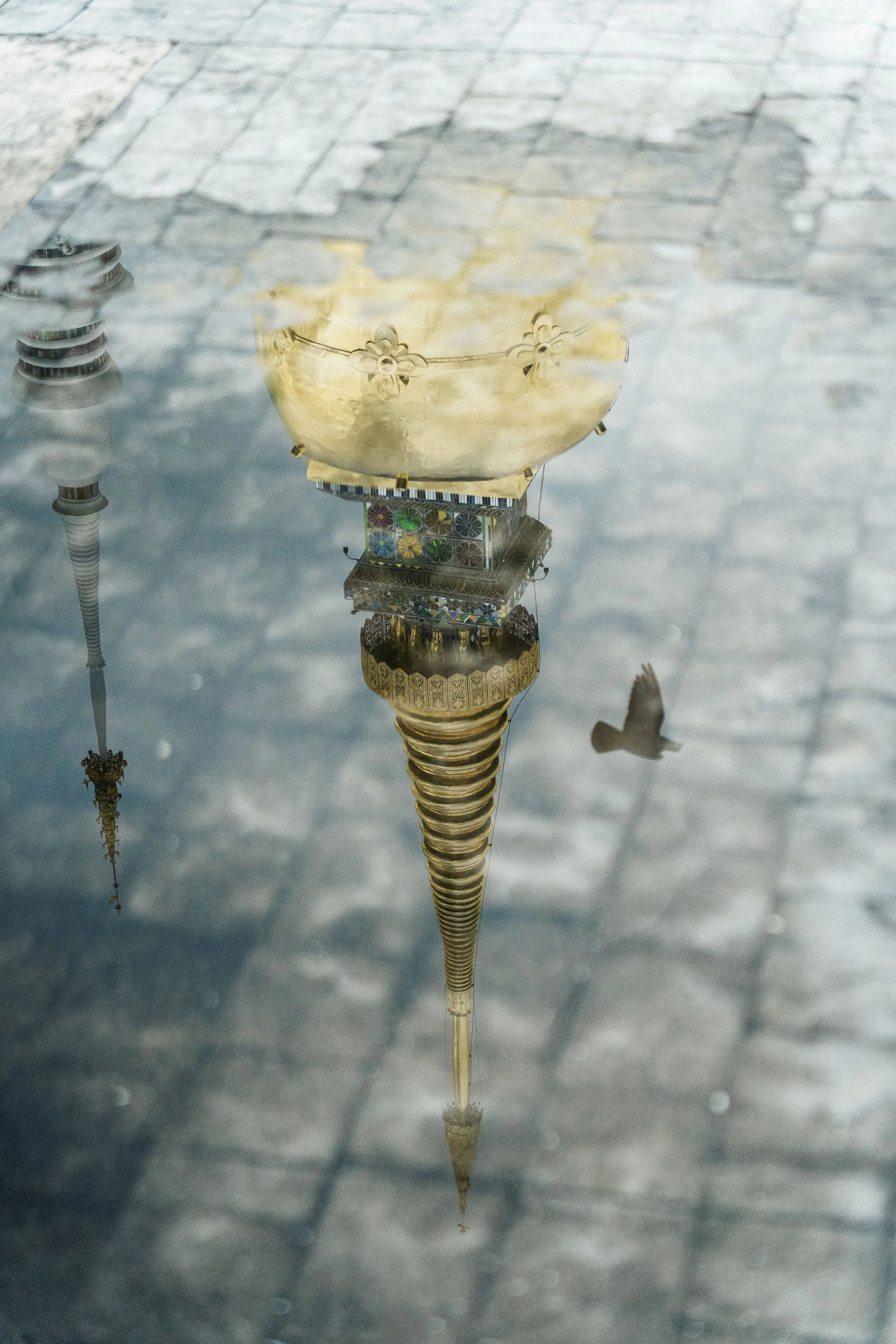Reflection of a tall ornate building with a pointed spire and smaller domes in a puddle, with a bird flying nearby.