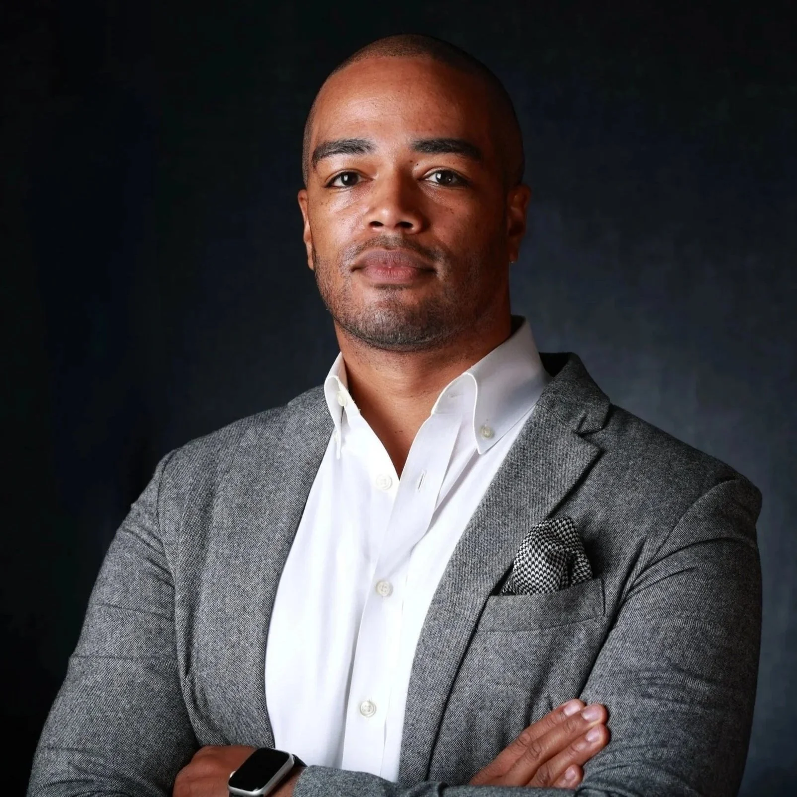 A man wearing a gray suit with a pocket square, white shirt, and an Apple Watch, posing with crossed arms against a dark background.