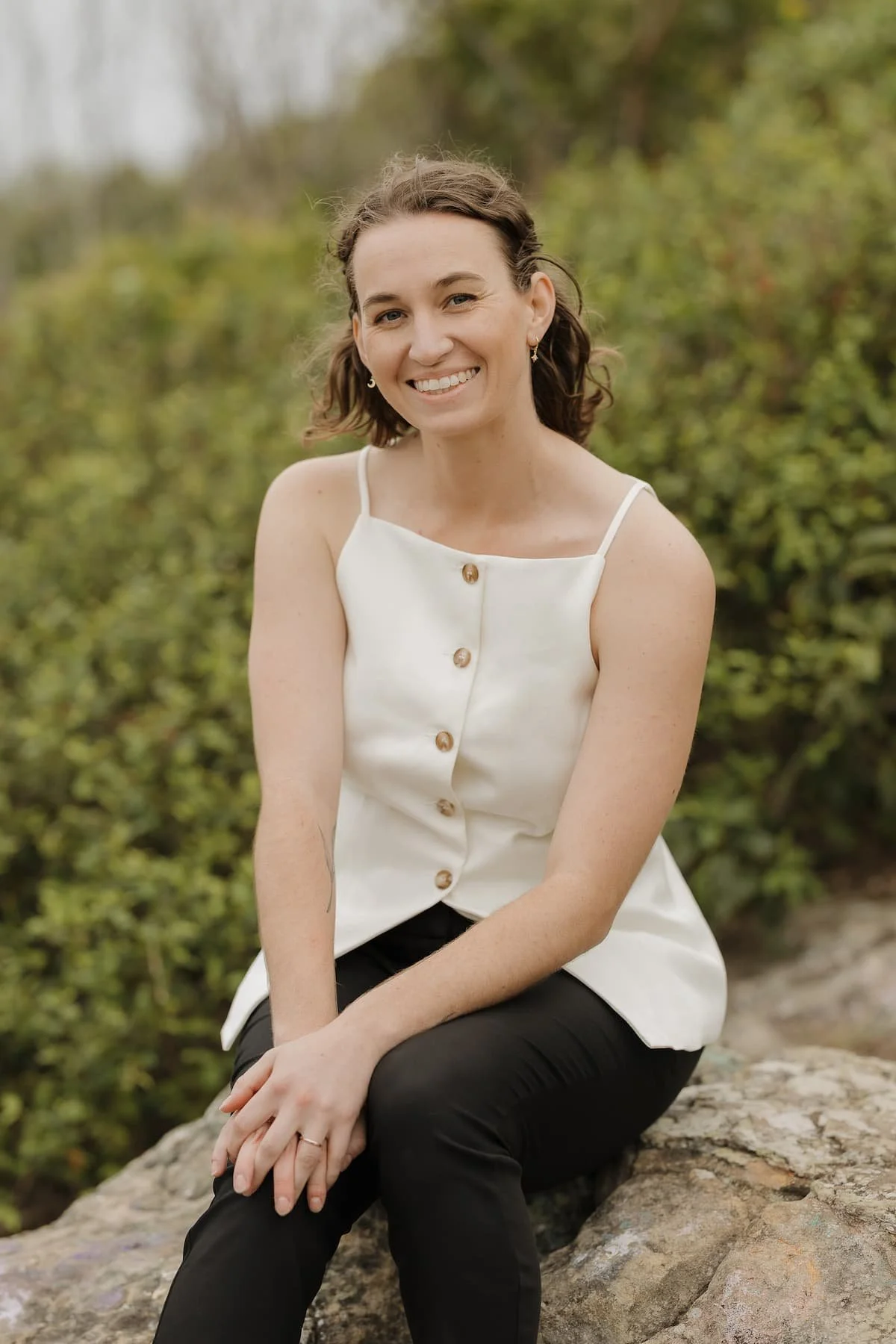 A woman with shoulder-length brown hair, smiling, wearing a white sleeveless button-down top and black pants, sitting outdoors on a large rock with green foliage in the background.