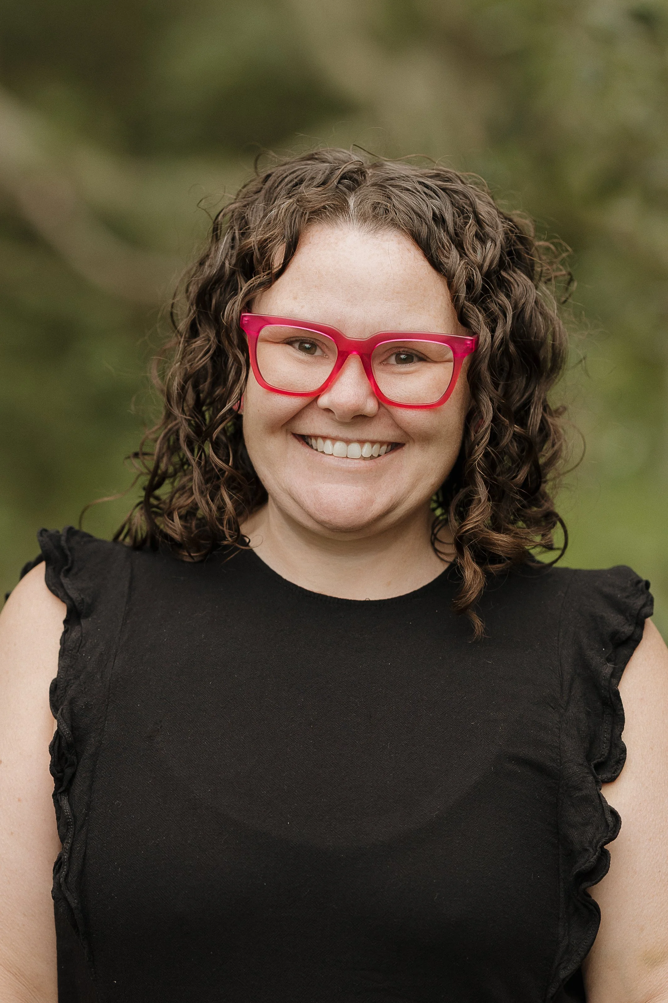 A woman with curly brown hair, wearing red glasses and a black top, smiling outdoors with a blurred green background.