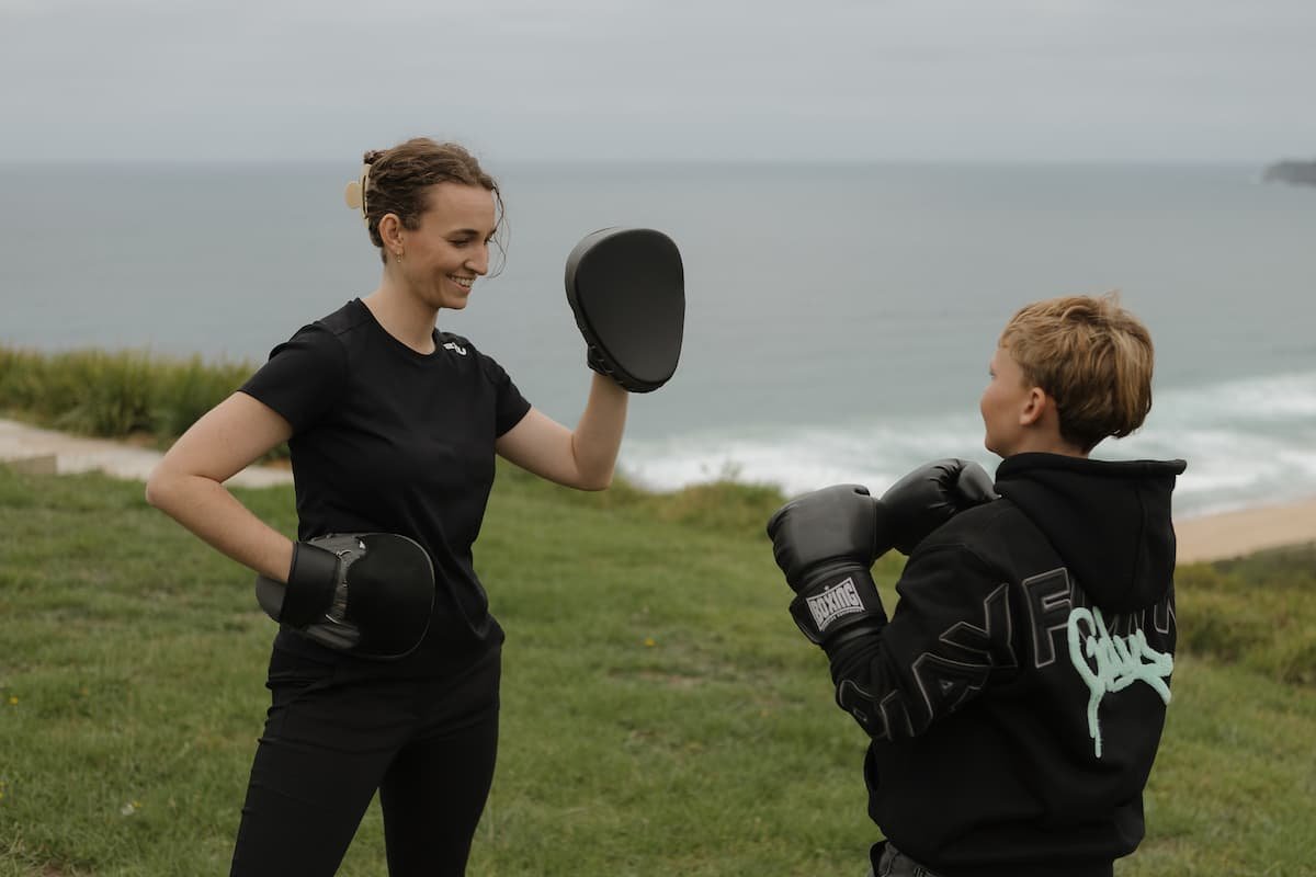 A woman and a boy practicing boxing outdoors near a body of water, with the woman holding focus mitts and the boy wearing boxing gloves.