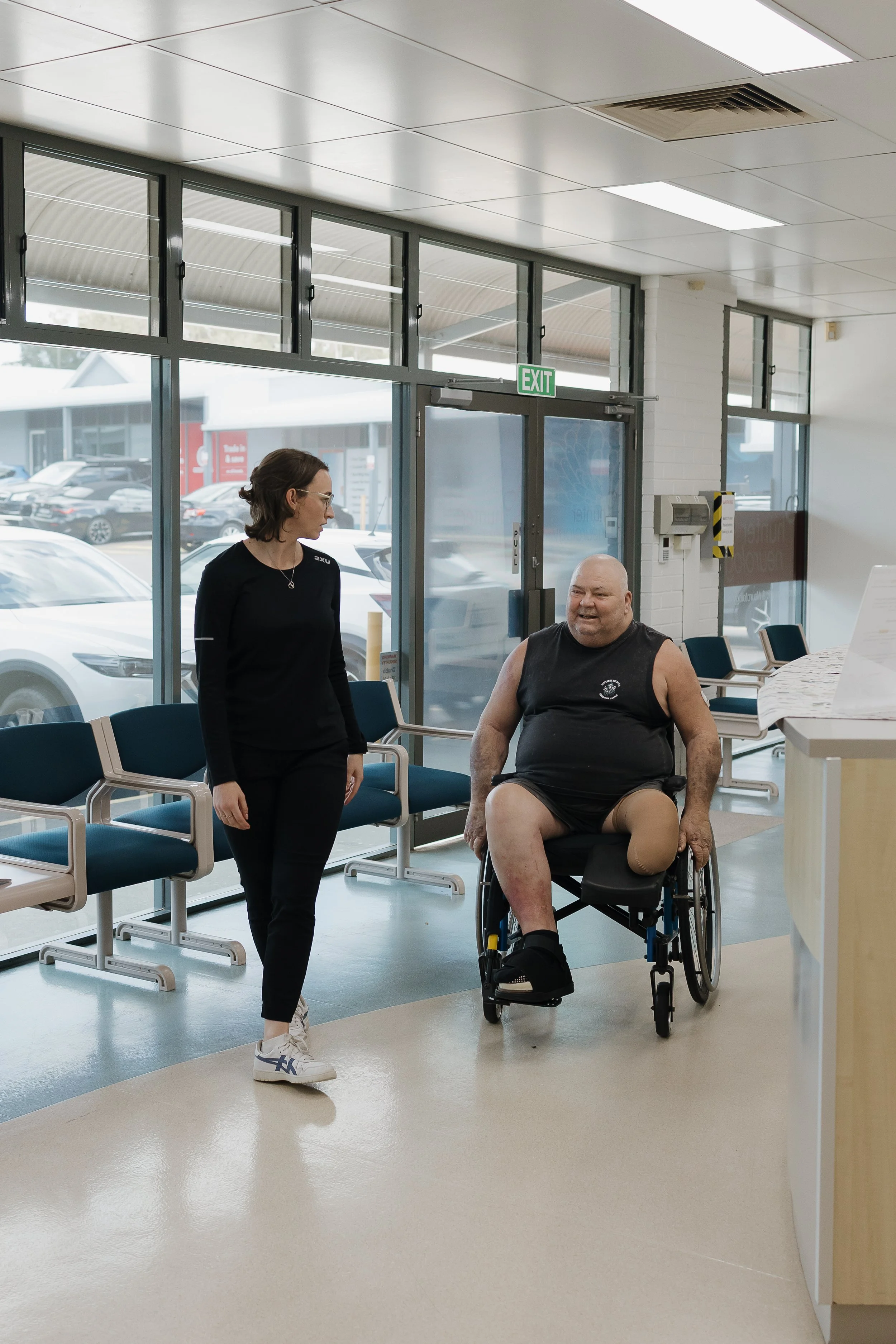 A woman and a man in a wheelchair at an airport waiting area, with large windows showing parked cars outside and an exit sign overhead.