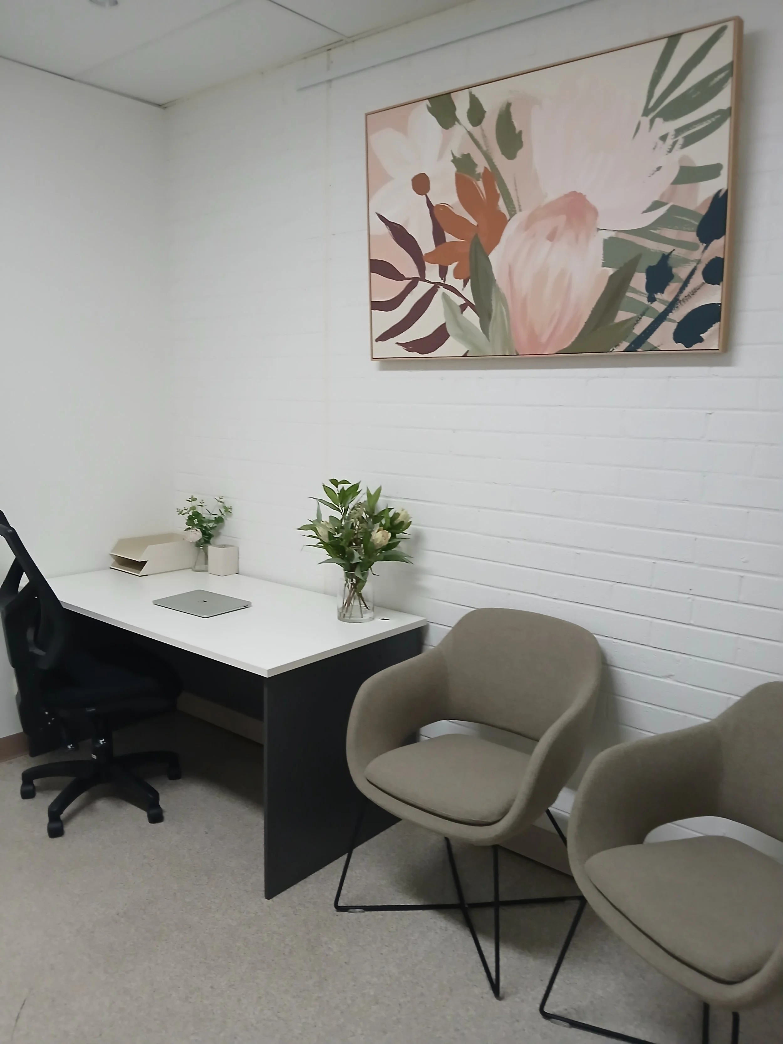 An office with a white wall, a white desk with a laptop and plants, a black office chair, two beige armchairs, and a large floral painting hanging on the wall.