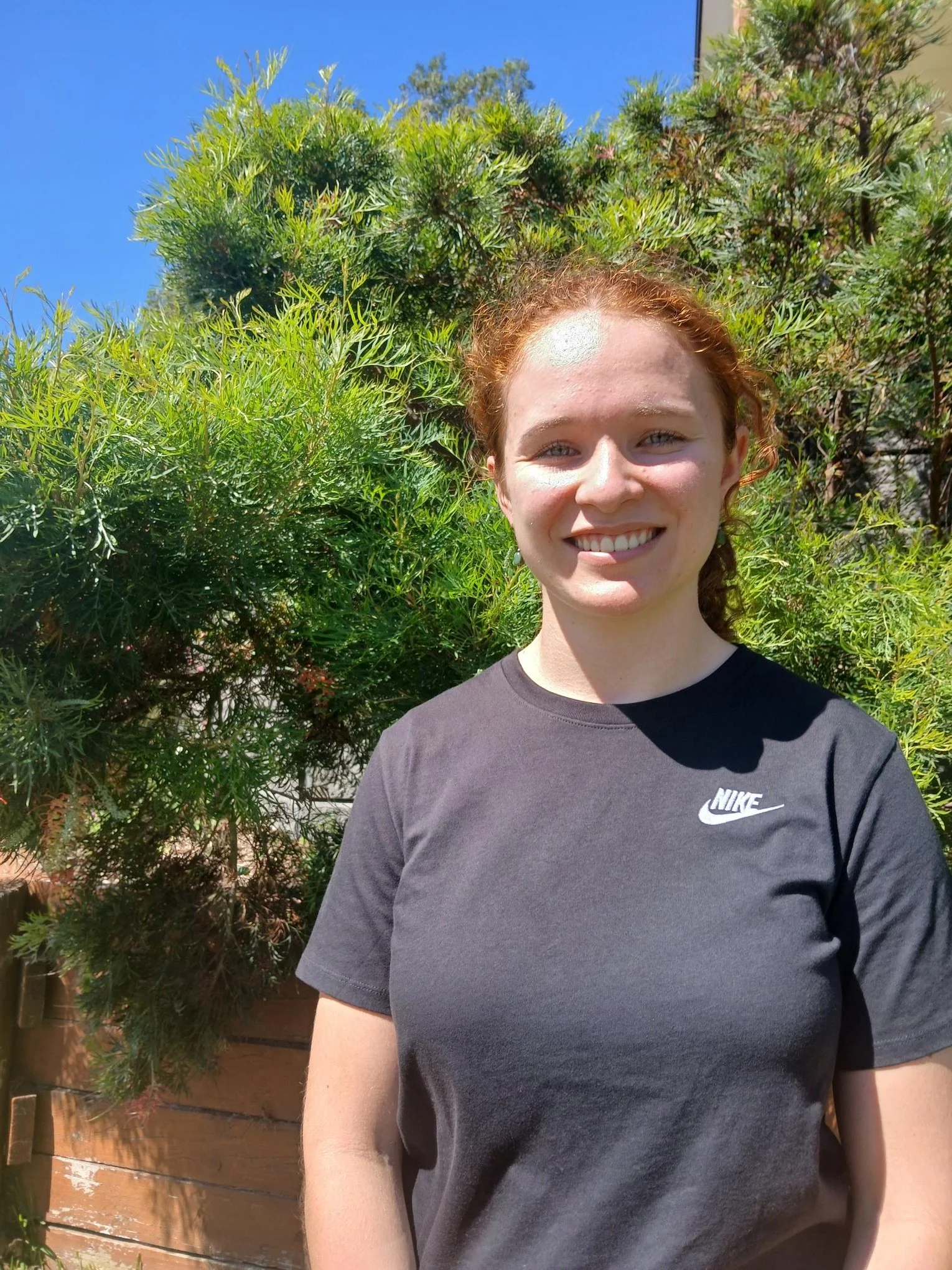 A young woman with red hair and a black Nike t-shirt smiling outdoors with green bushes and a clear blue sky in the background.