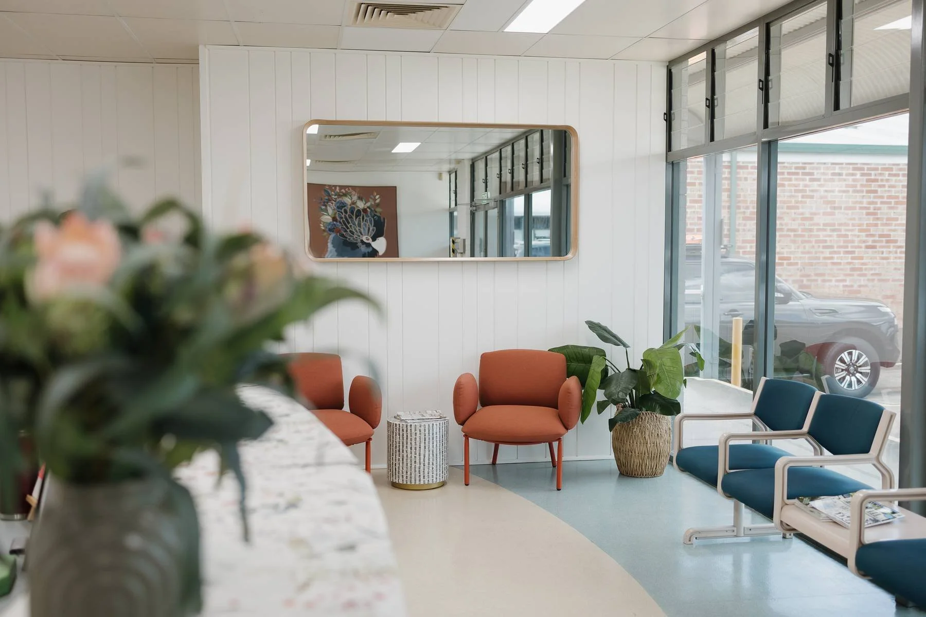 Waiting area in a modern office or clinic with orange and blue chairs, potted plants, a mirror on the wall, and large windows showing a brick building and parked car outside.