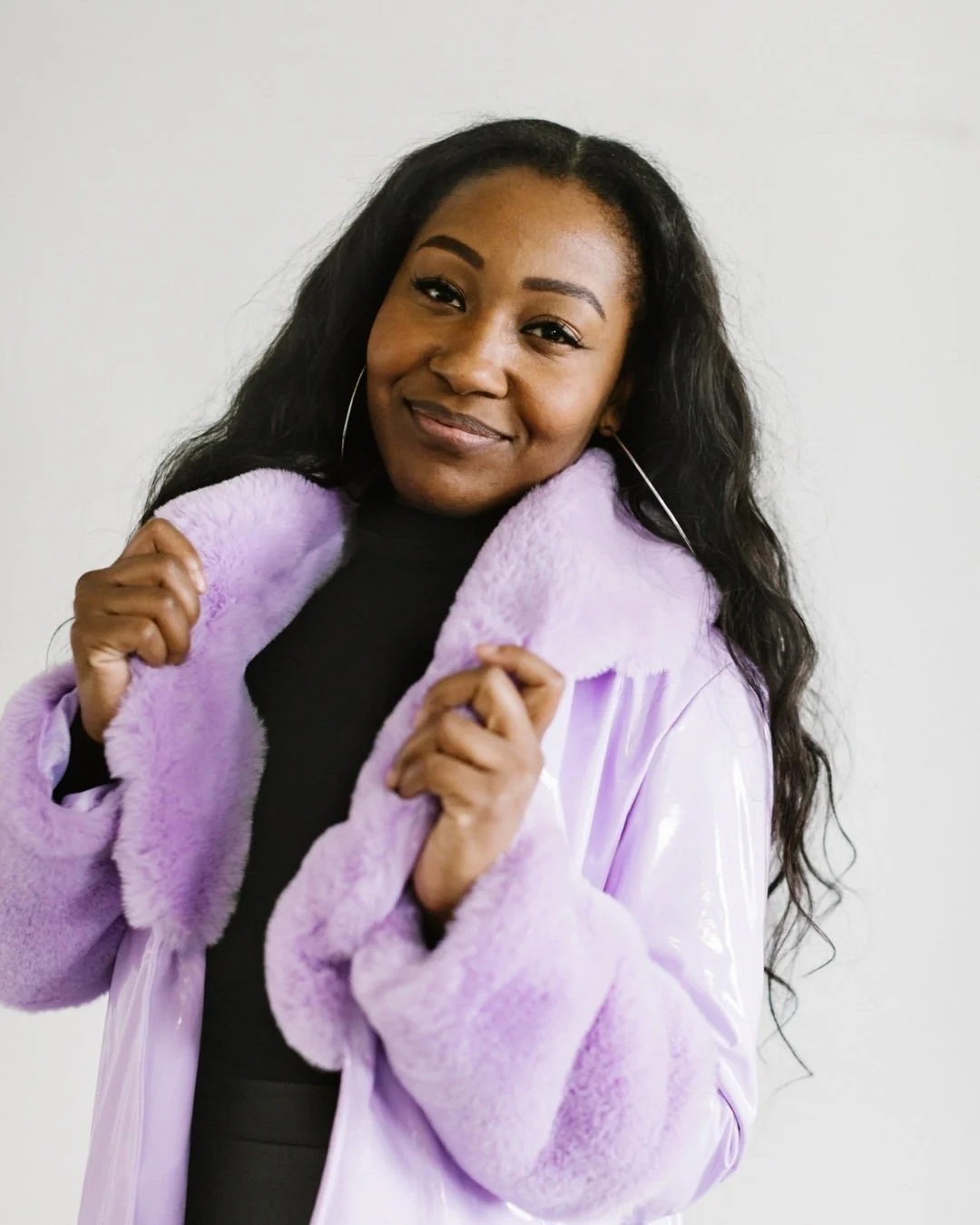 A woman with long dark hair is smiling at the camera, wearing a black top and a lavender-colored fur-lined jacket.