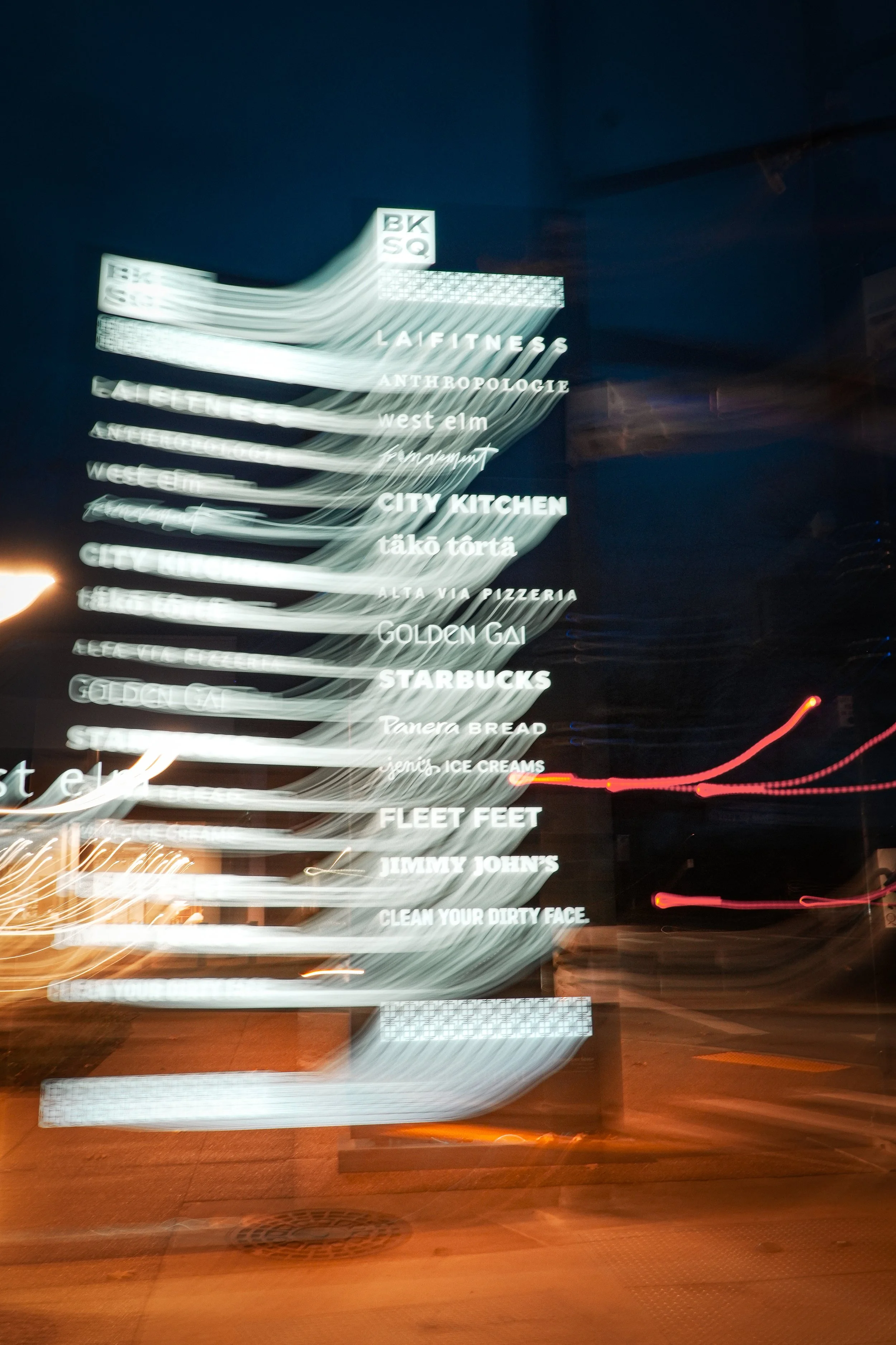 Blurry illuminated directory sign at night listing various businesses including LA FITNESS, ANTHROPOLOGIE, west elm, CITY KITCHEN, GOLDEN GAI, STARBUCKS, Panera BREAD, JIMMY JOHN'S, CLEAN YOUR DIRTY FACE, and others.