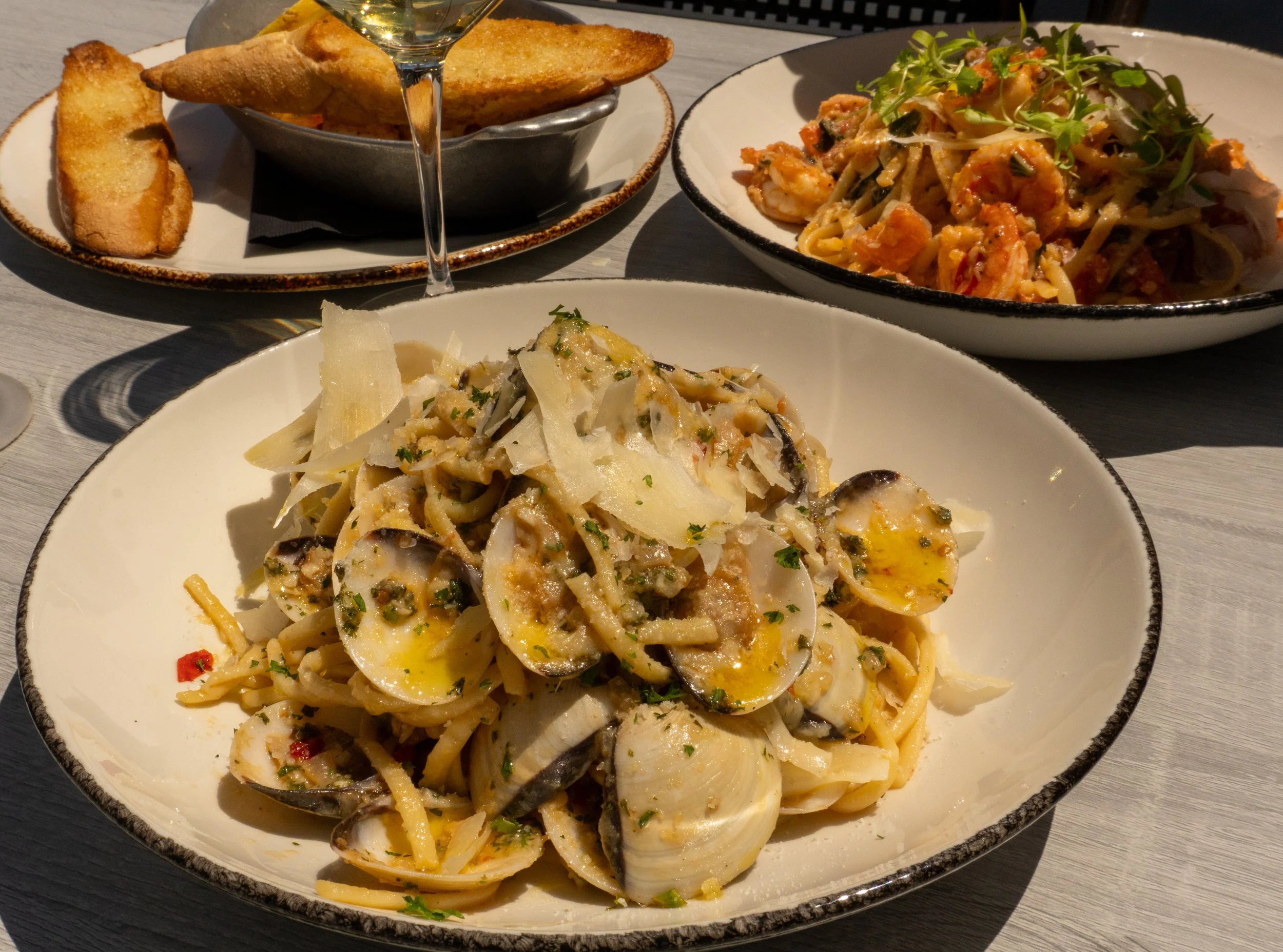 A plate of pasta with clams, shaved cheese, and chopped herbs in the foreground, a bowl of toasted bread and a glass of white wine behind, and a bowl of seafood pasta with microgreens on top in the background.