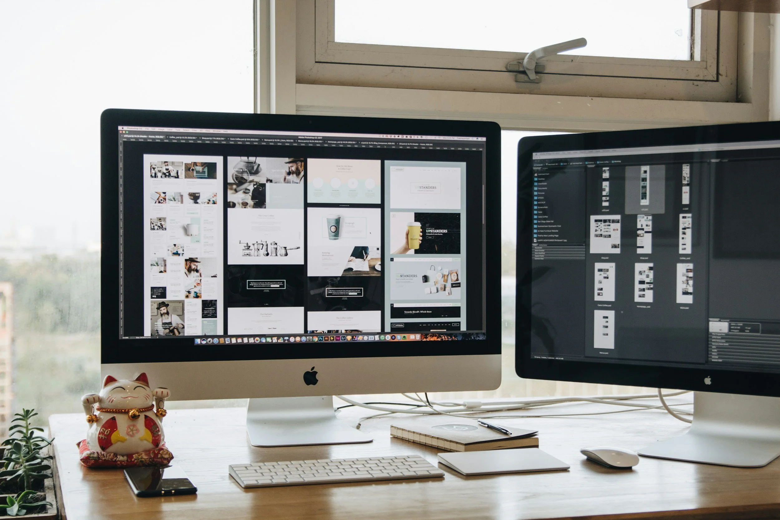 Two Apple iMac computers on a wooden desk with design folders, a notebook, a pen, a wireless keyboard, a mouse, a smartphone, and a Maneki-neko statue, with a window in the background.