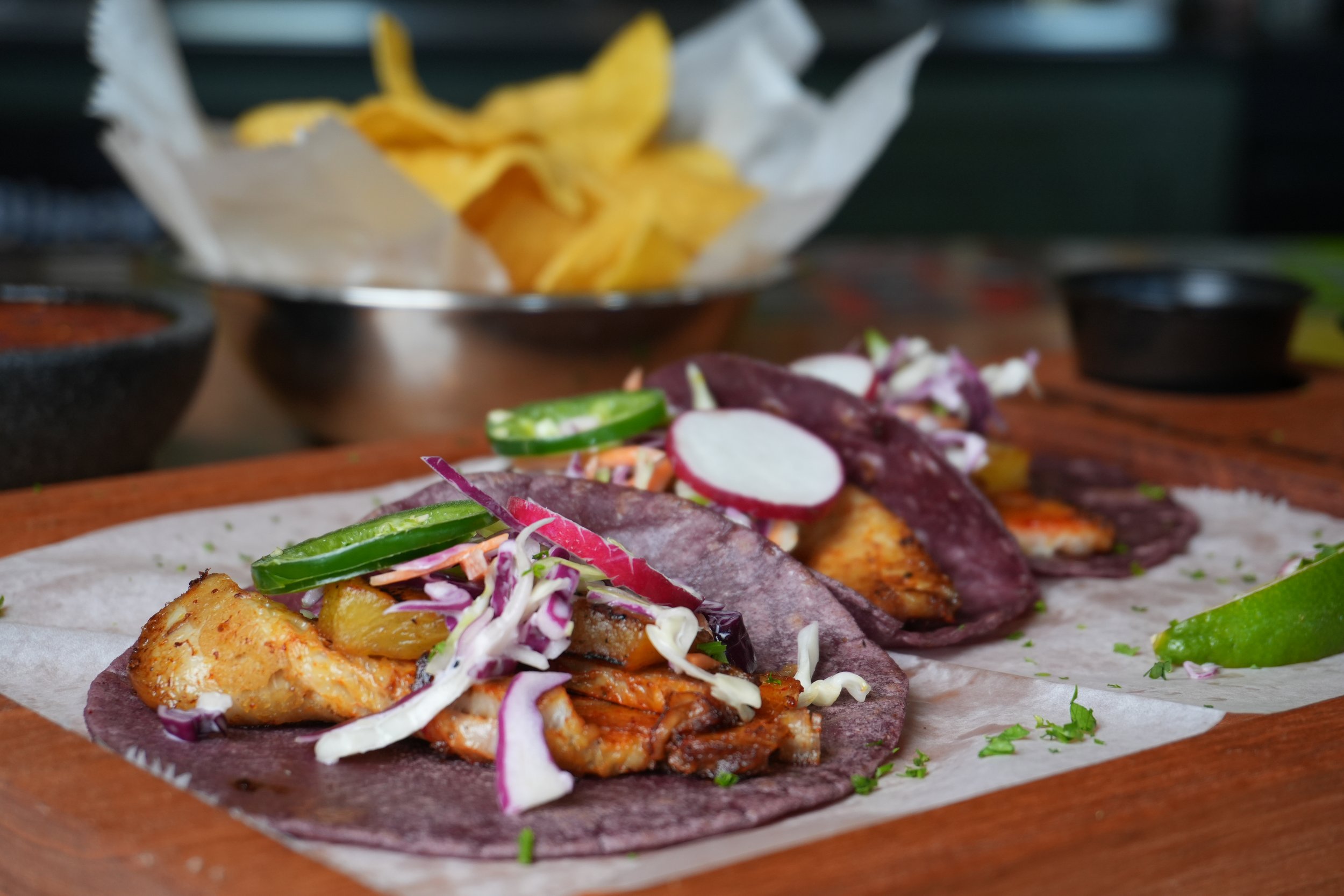 A close-up of three fish tacos topped with sliced cucumbers, radishes, shredded cabbage, and grilled fish, served on a wooden platter with a lime wedge.