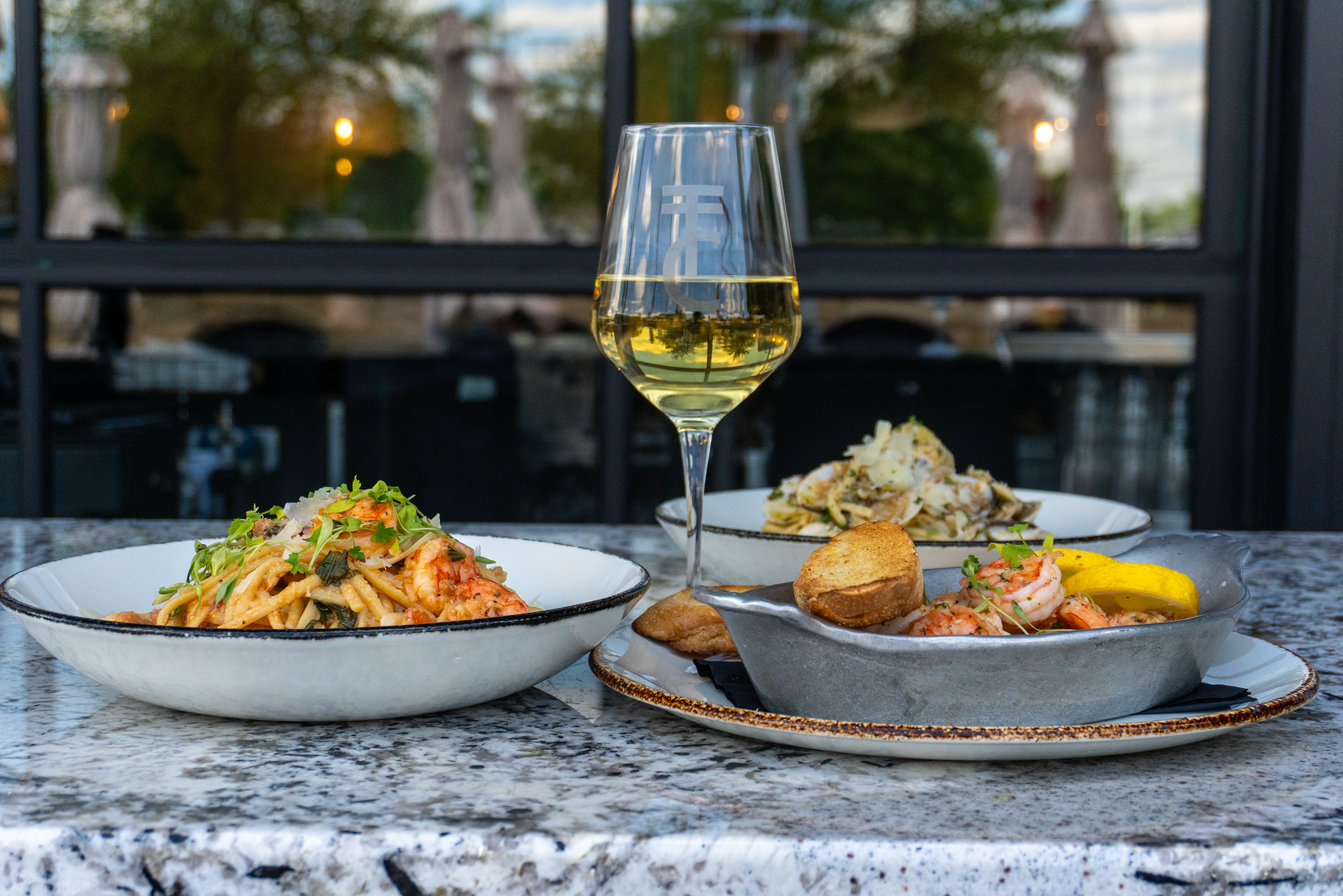 A glass of white wine on a table with two plates of pasta, one with shrimp and lemon garnishes, and the other with seafood, served on a granite table outside with trees in the background.