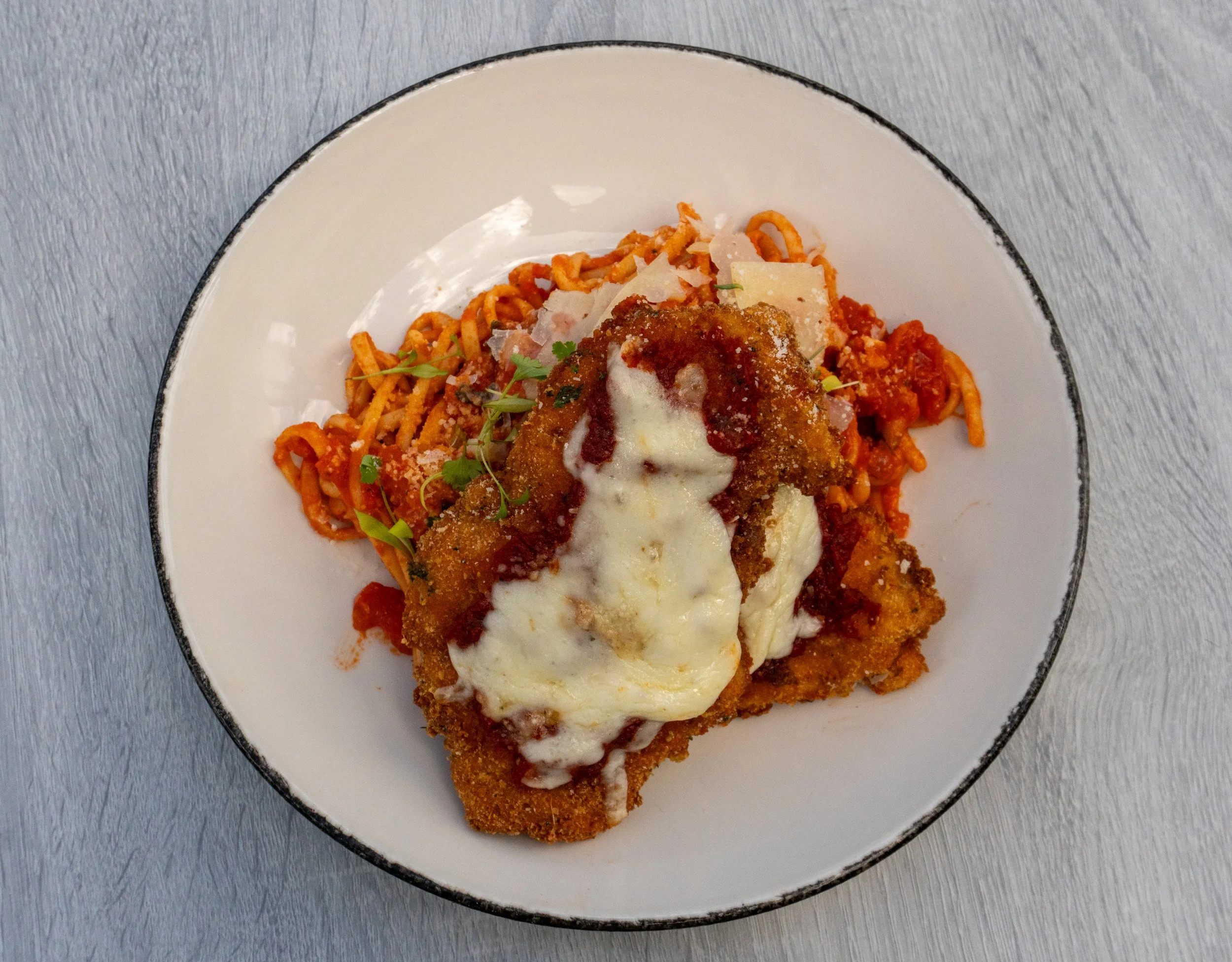 A plate of spaghetti with marinara sauce topped with breaded chicken, melted mozzarella, and fresh herbs, served on a white dish with a black rim.