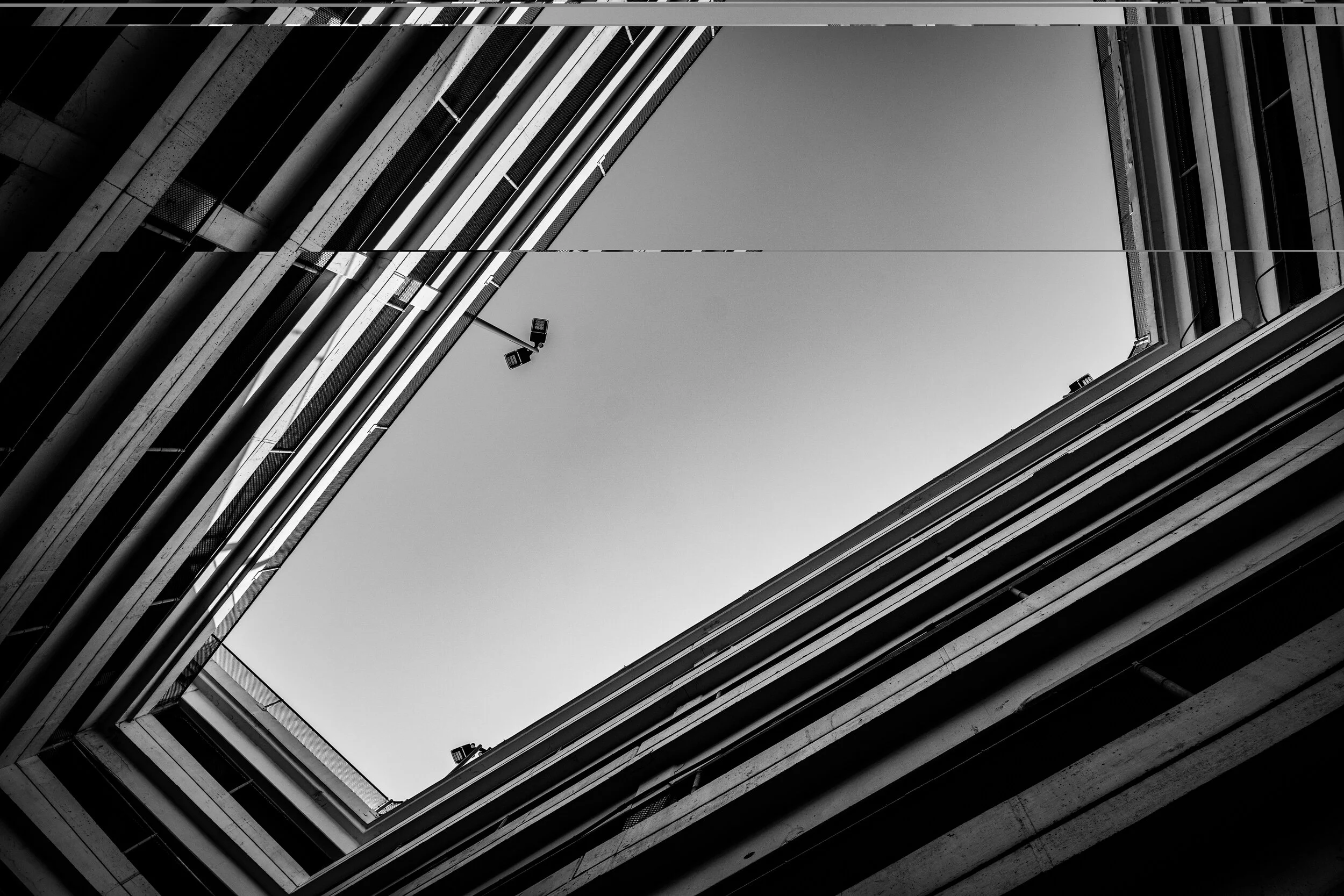 Looking up between tall concrete buildings, showing a square opening with a clear sky and security cameras mounted on the edges, in black and white.