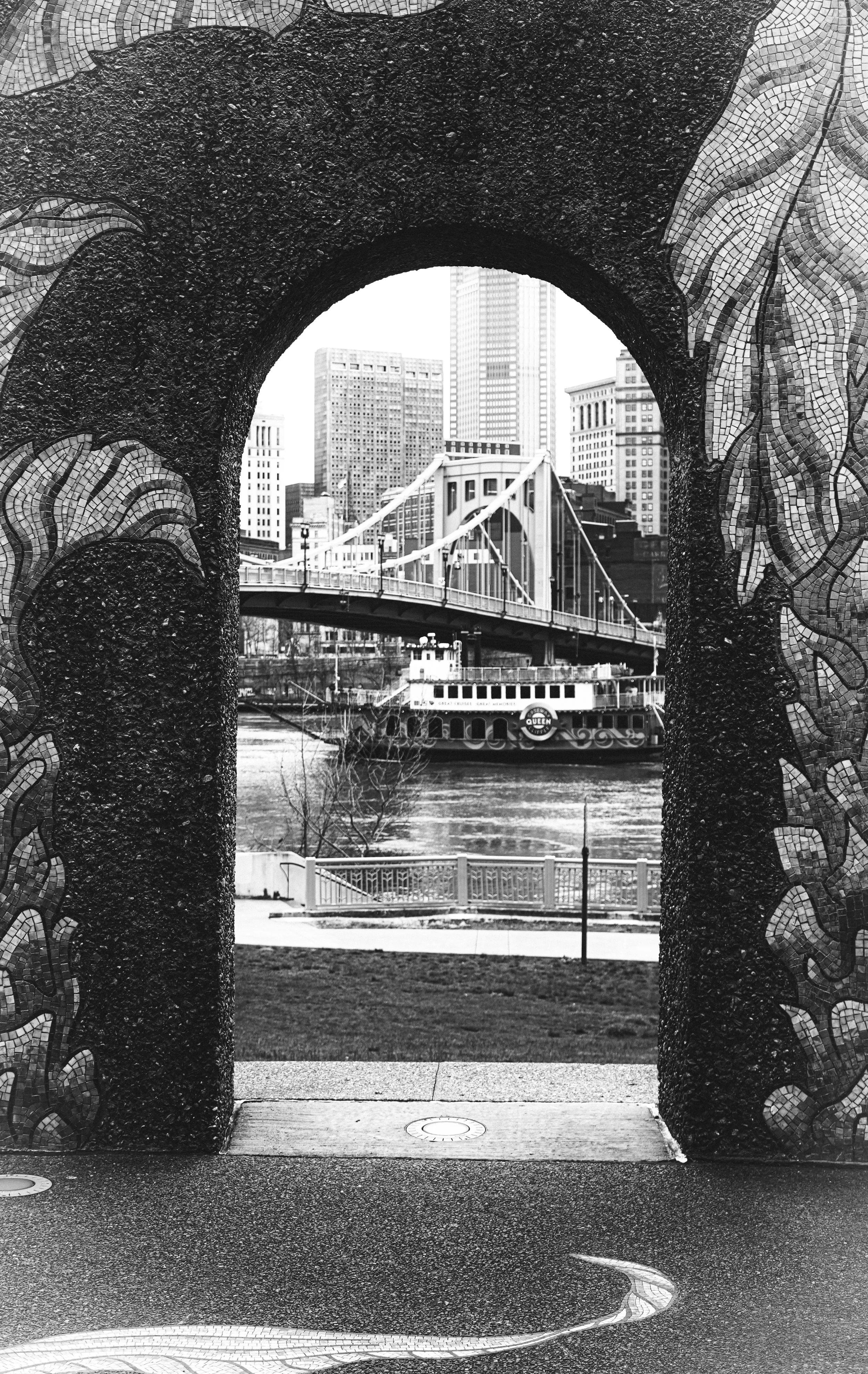 View through a decorative archway revealing a cityscape with a suspension bridge over a river, surrounded by tall buildings in the background.