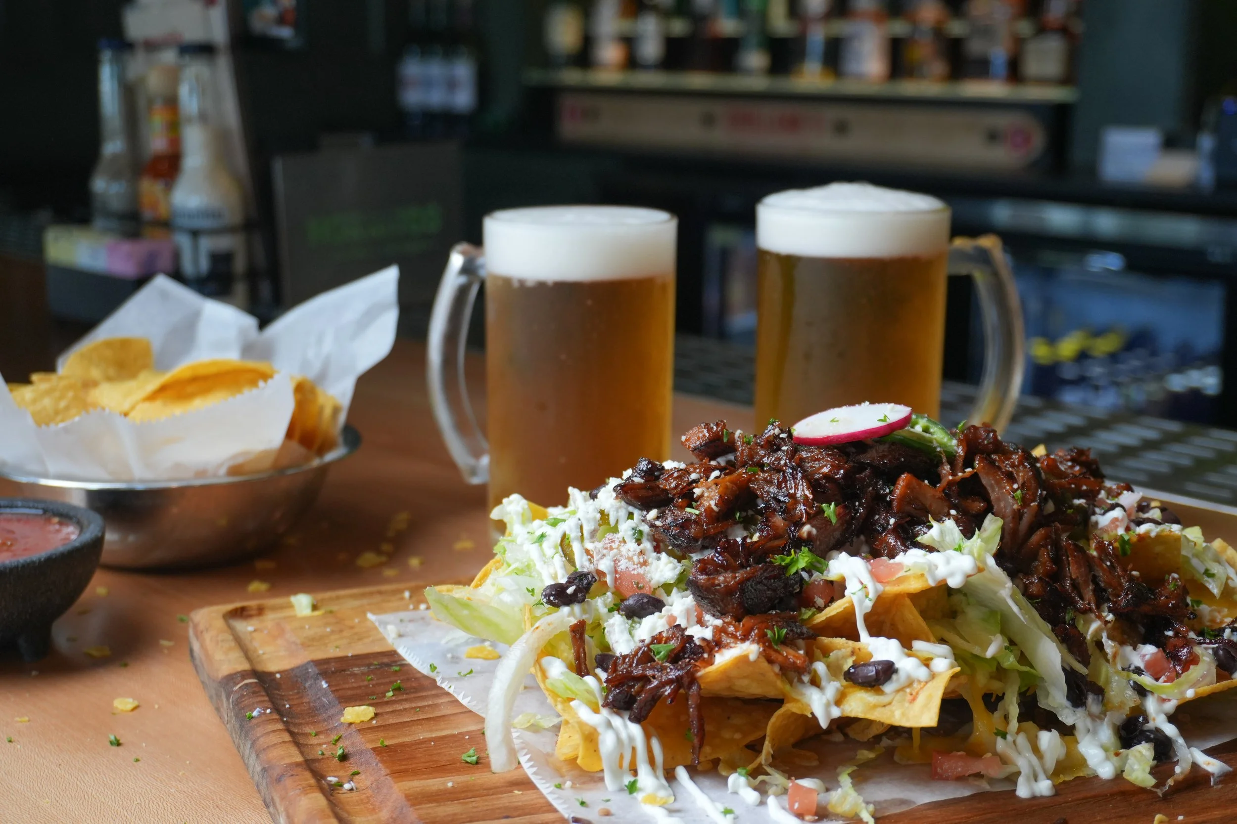 Two mugs of beer, nachos with shredded lettuce, cheese, black beans, shredded meat, and vegetables, a bowl of potato chips, and a bowl of salsa in a restaurant or bar setting.