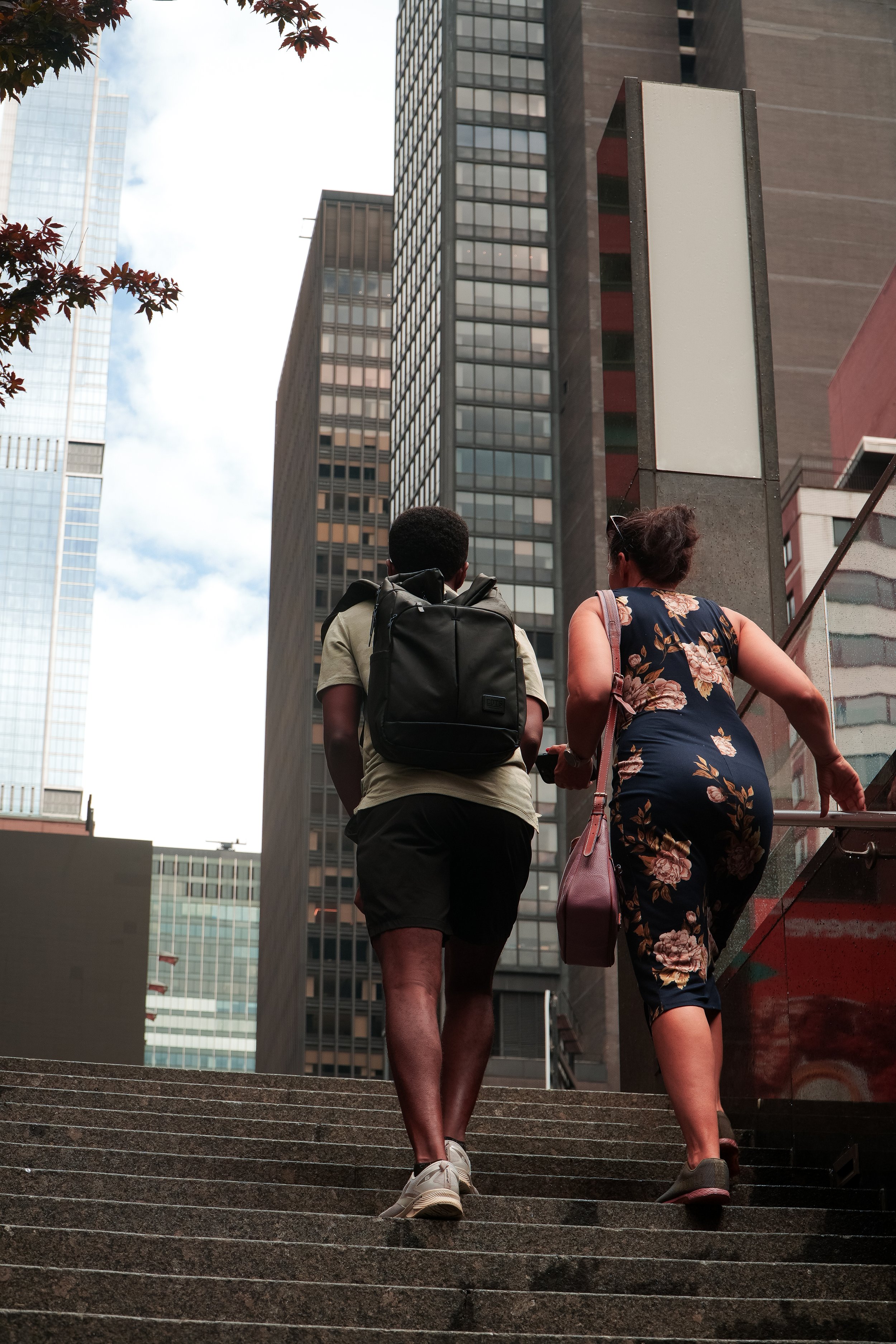 A man and woman walking up stairs in a city, surrounded by tall office buildings, with a cloudy sky above.