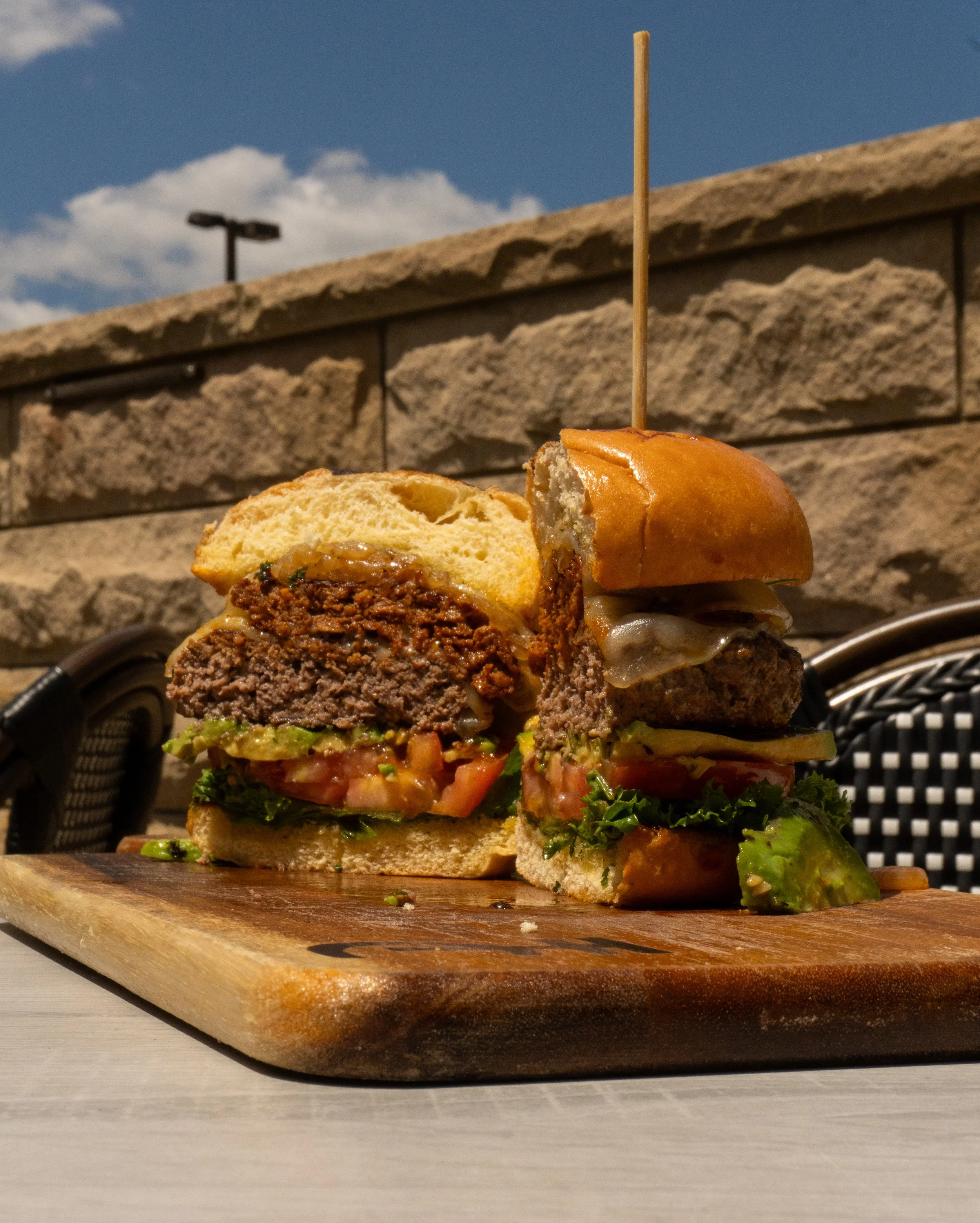 A cross-section of a cheeseburger with lettuce, tomato, cheese, and a beef patty, placed on a wooden cutting board outdoors, with a stone wall and blue sky in the background.