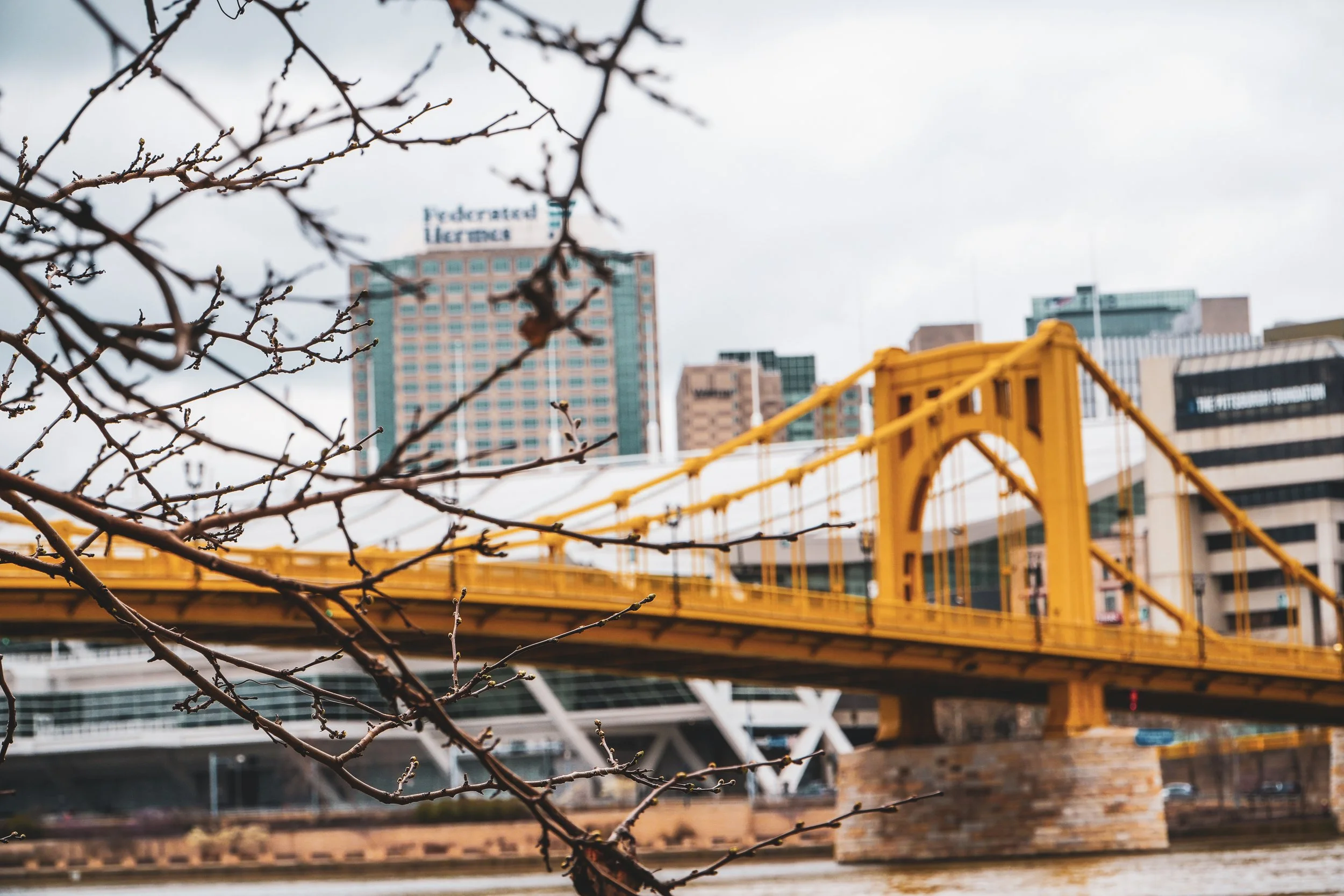 A yellow bridge extending over a river with downtown buildings in the background.