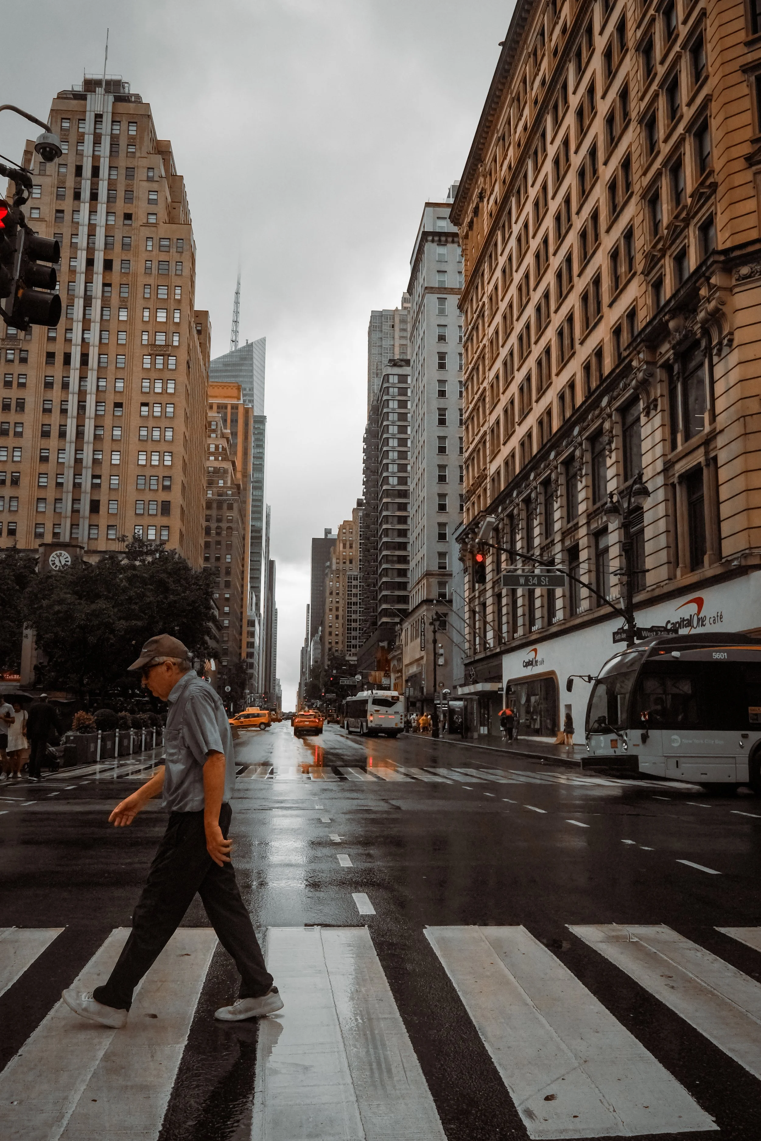 A person walking across a wet crosswalk on a city street with tall buildings on either side, and a cloudy sky above.