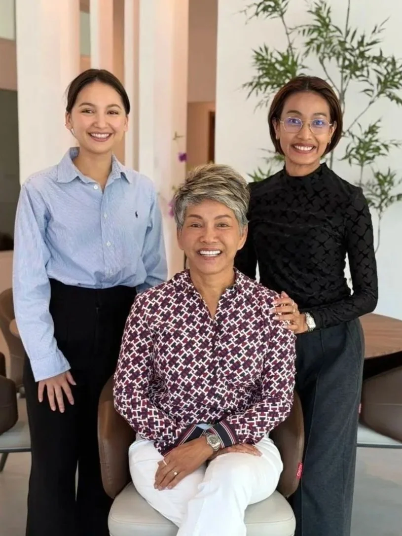 Three women smiling indoors, one seated and two standing behind her, with plants and neutral decor in the background.