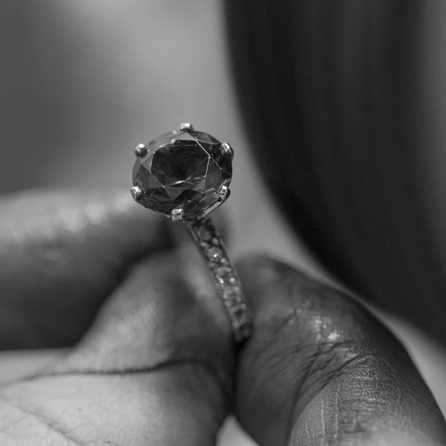 A close-up black and white photograph of a person's hand holding a ring with a large, round gemstone.