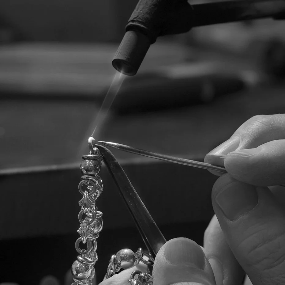 A person cleaning a silver chain with tools and a spray, close-up in black and white.