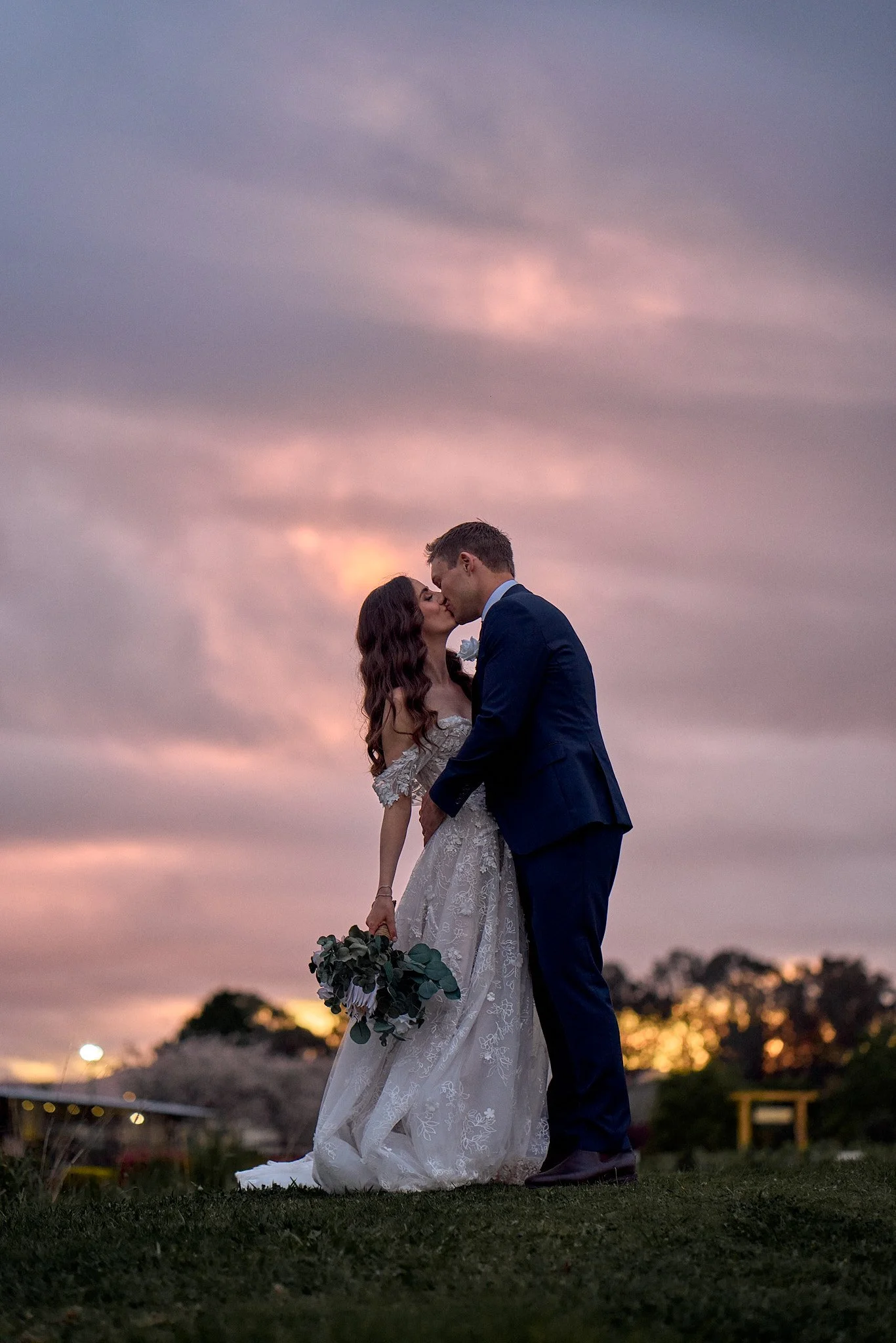 Bride and groom kissing at sunset, holding bouquet.