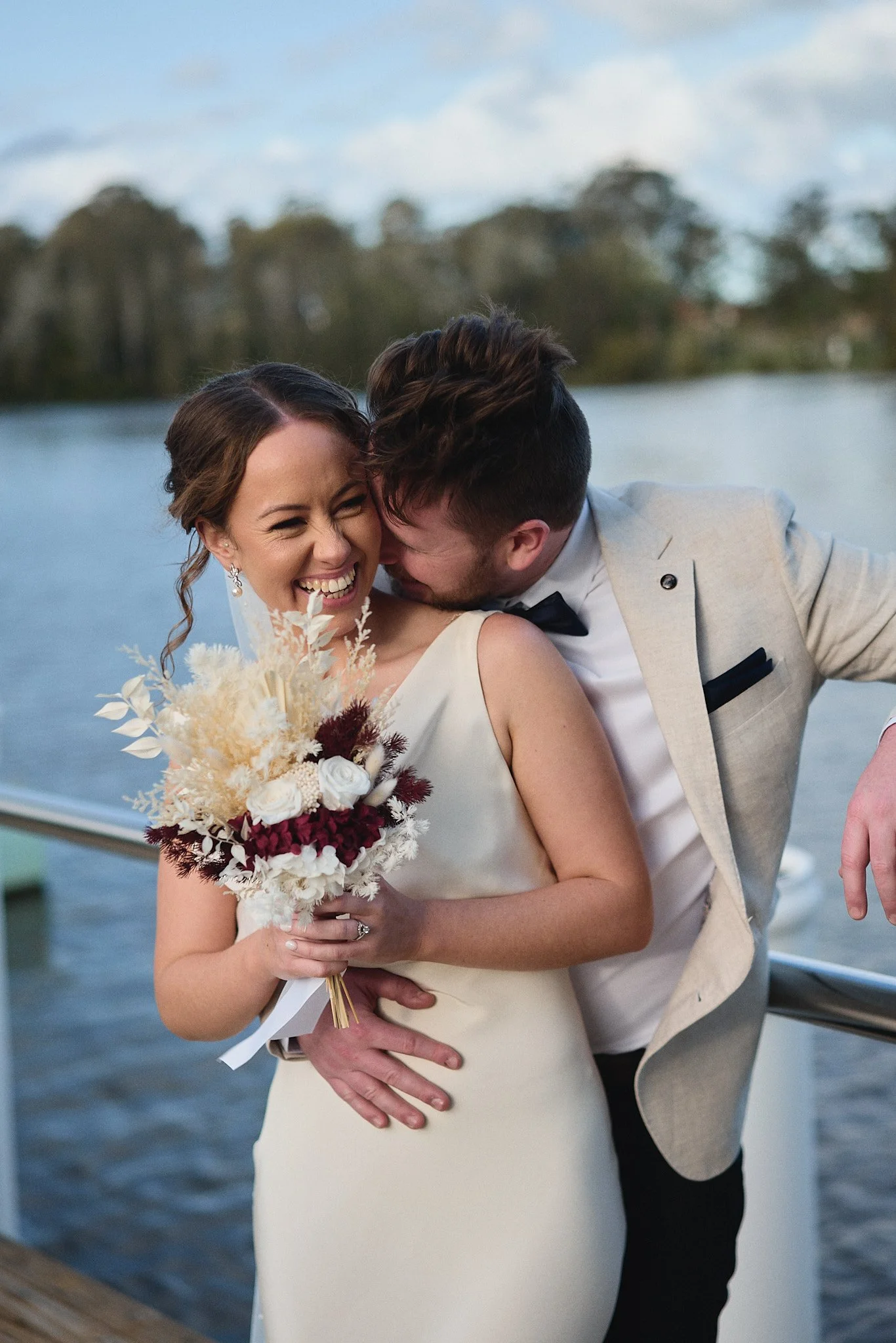Happy couple embracing by a lake, bride in white dress holding bouquet, groom in light suit.