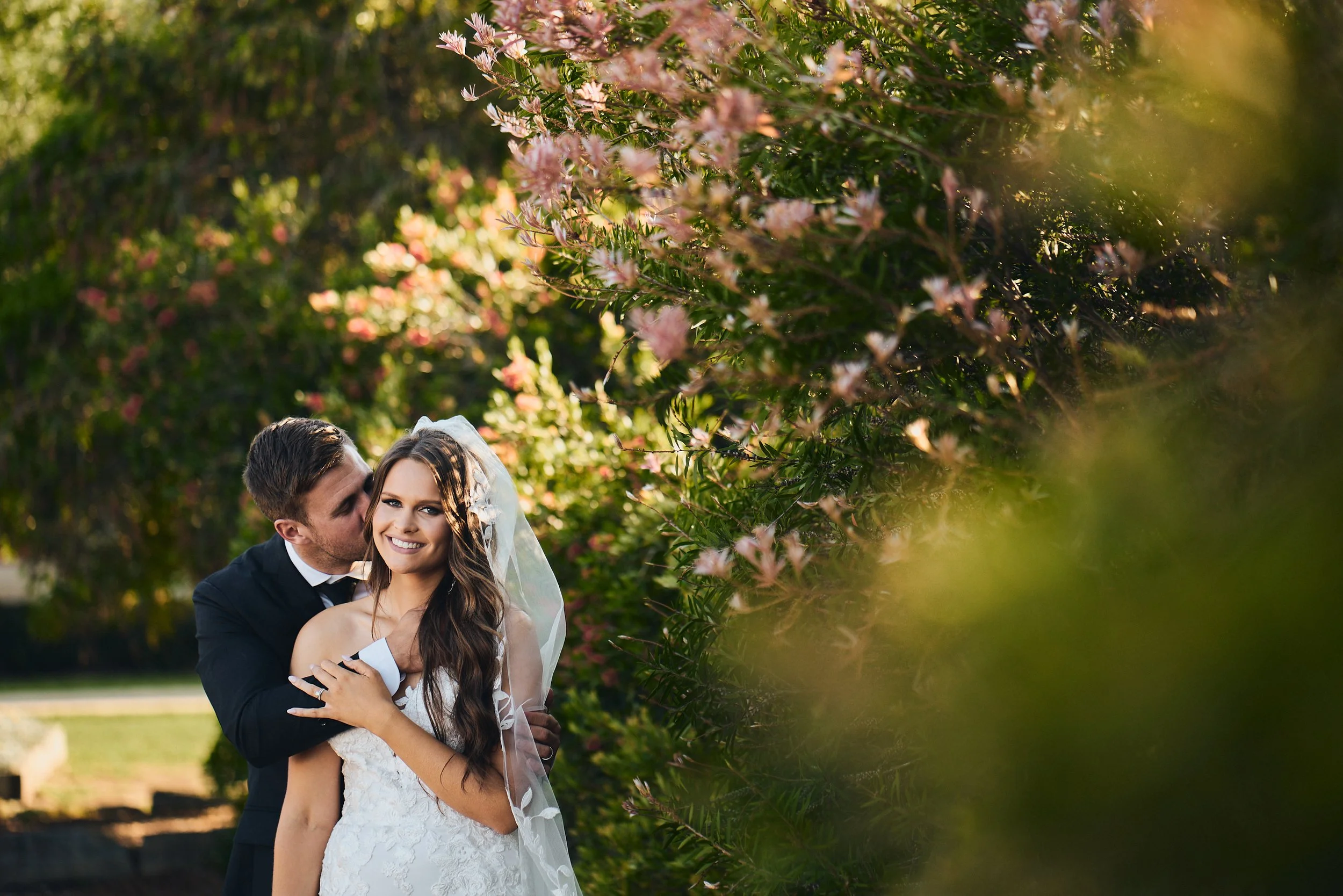 Bride and groom embracing outdoors with trees and flowers in background