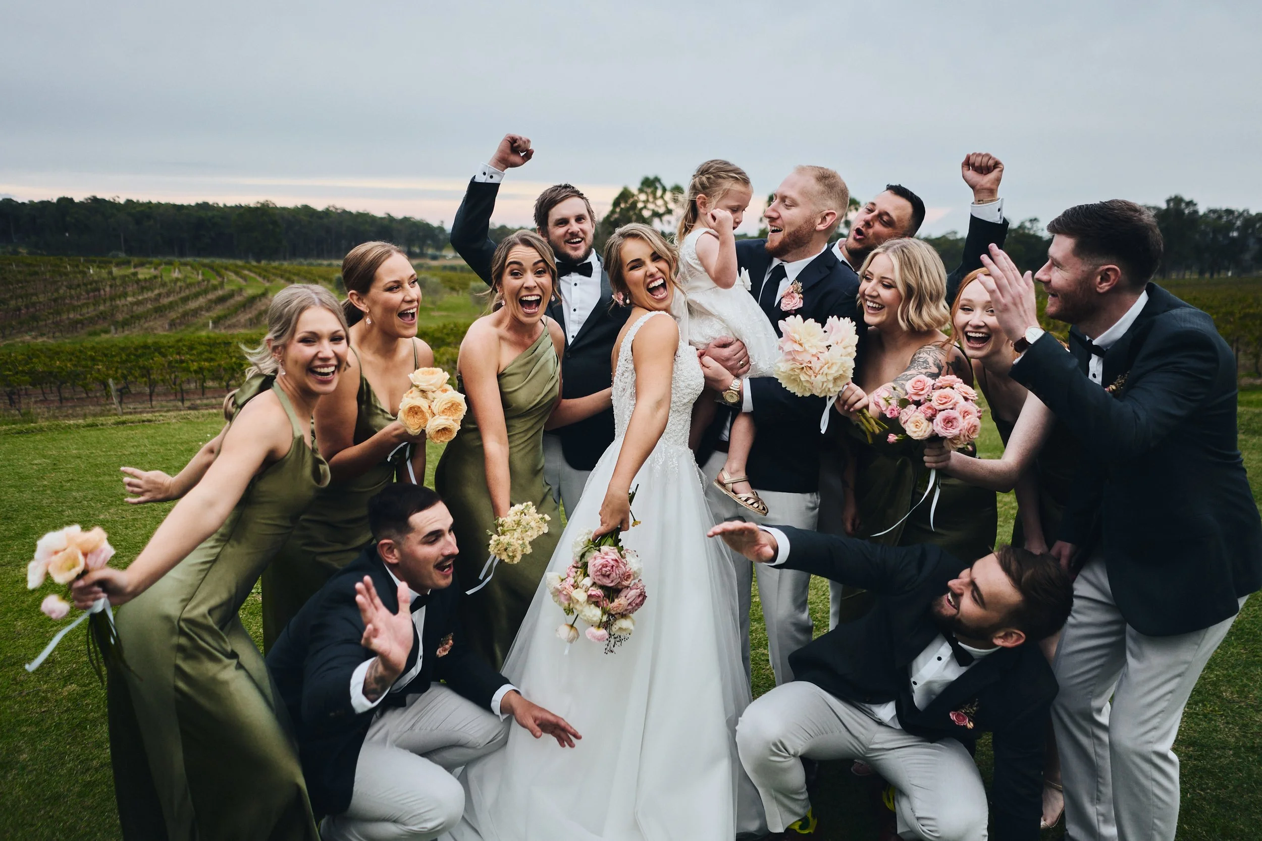 A joyful wedding party with bride, groom, bridesmaids in green dresses, and groomsmen in black suits with white pants. They're outdoors, holding floral bouquets, celebrating together. Vineyard backdrop.