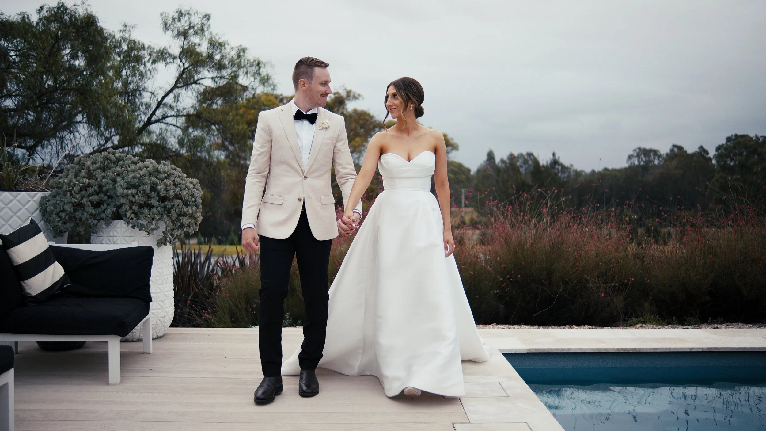 A bride and groom holding hands, walking along a patio next to a swimming pool, with trees and plants in the background.