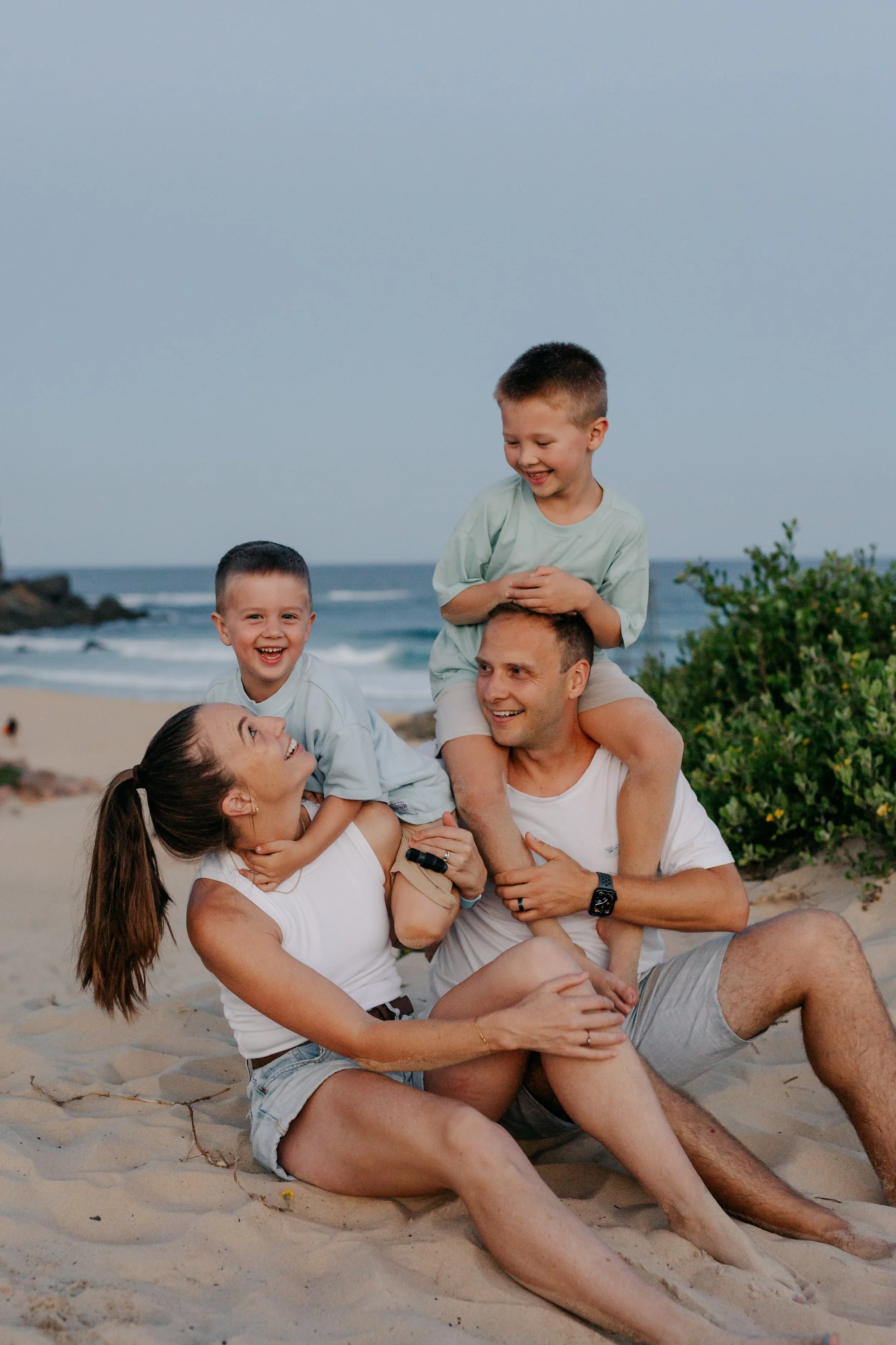 A family of four enjoying a day at the beach, with the father carrying a young boy on his shoulders while the mother holds a young girl on her lap, all smiling and laughing.