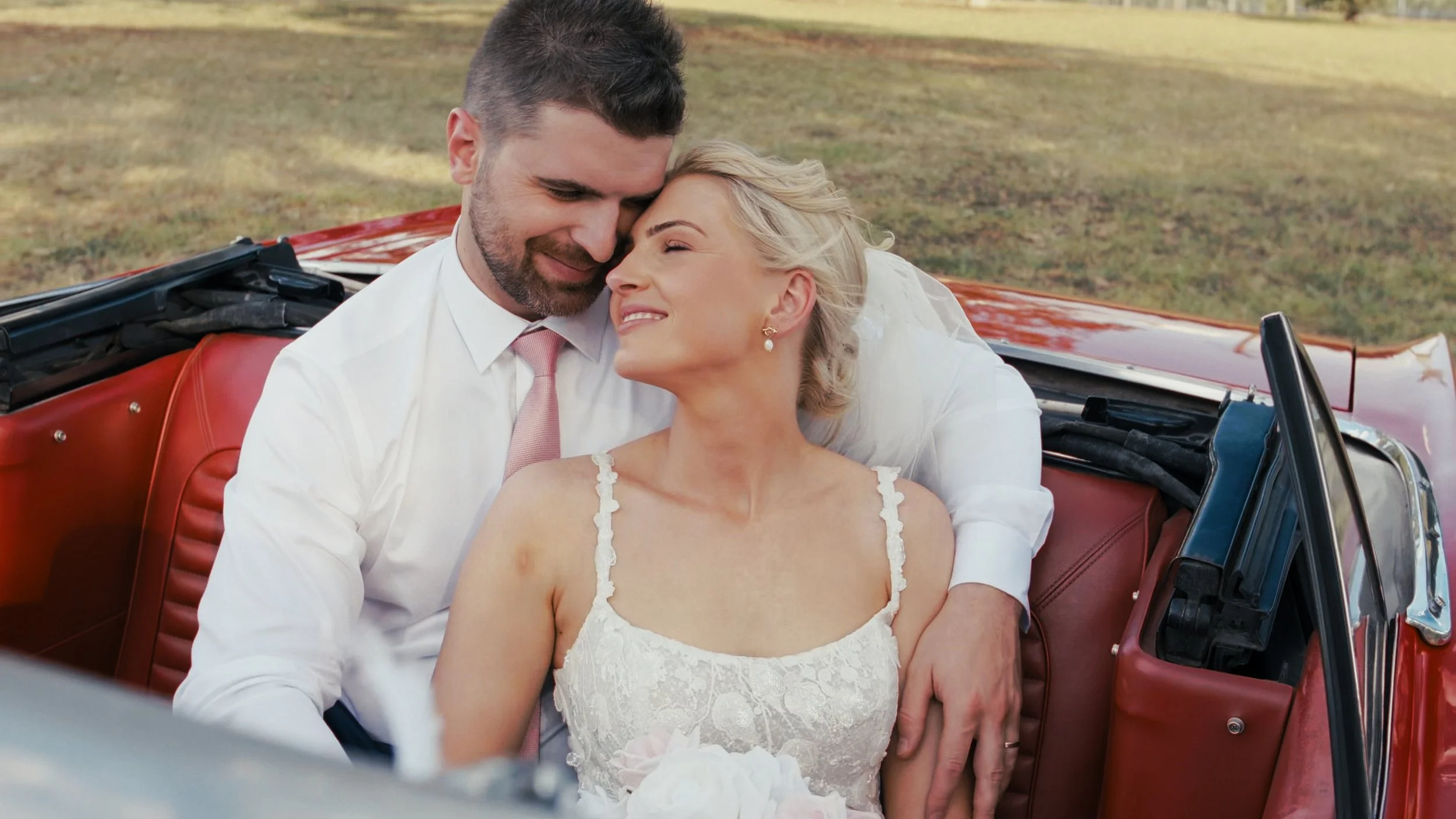 Smiling couple in a convertible car, bride in white dress, groom in white shirt and pink tie, outdoors.