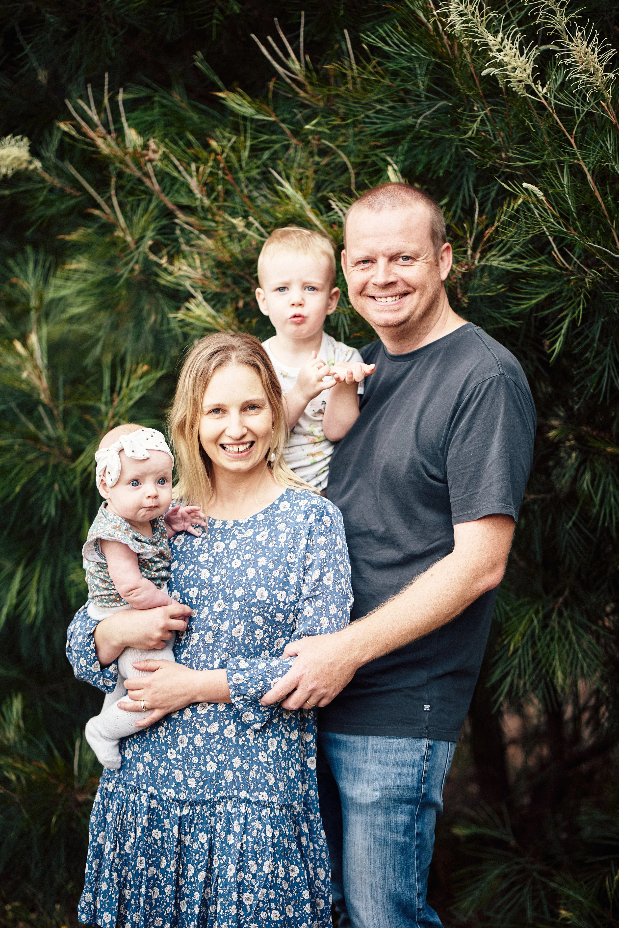 Family of four outdoors, smiling, with two young children and green foliage in the background.