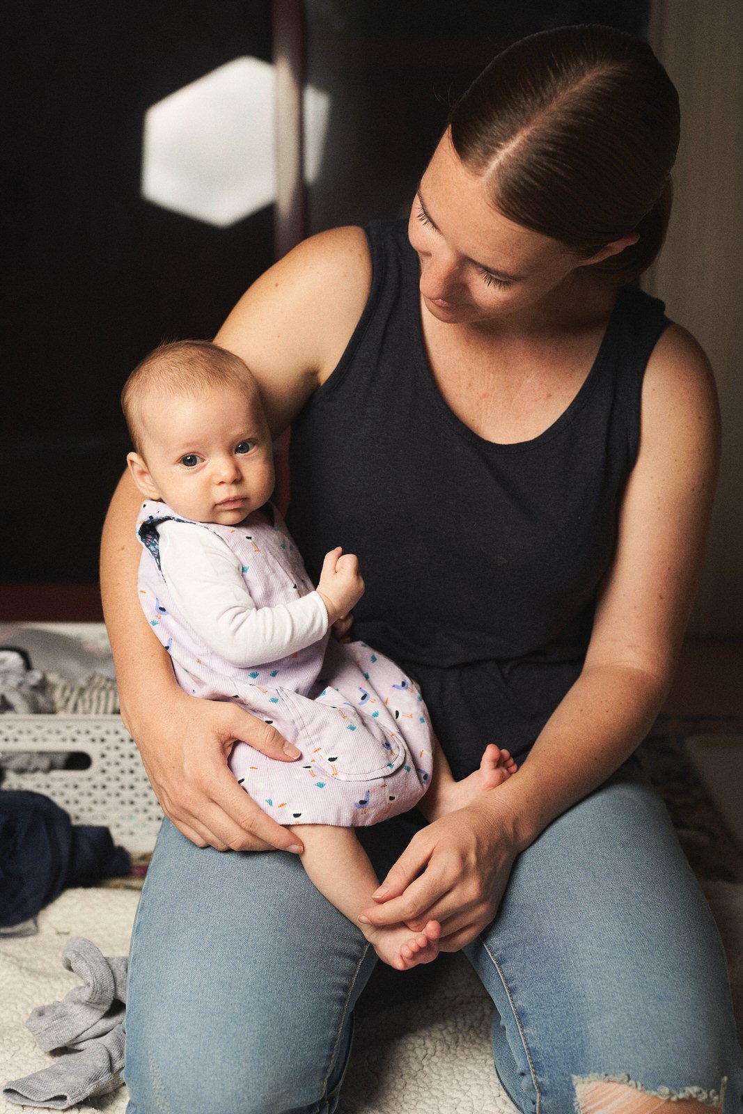 Mum holding a baby on lap inside a room, with laundry basket nearby