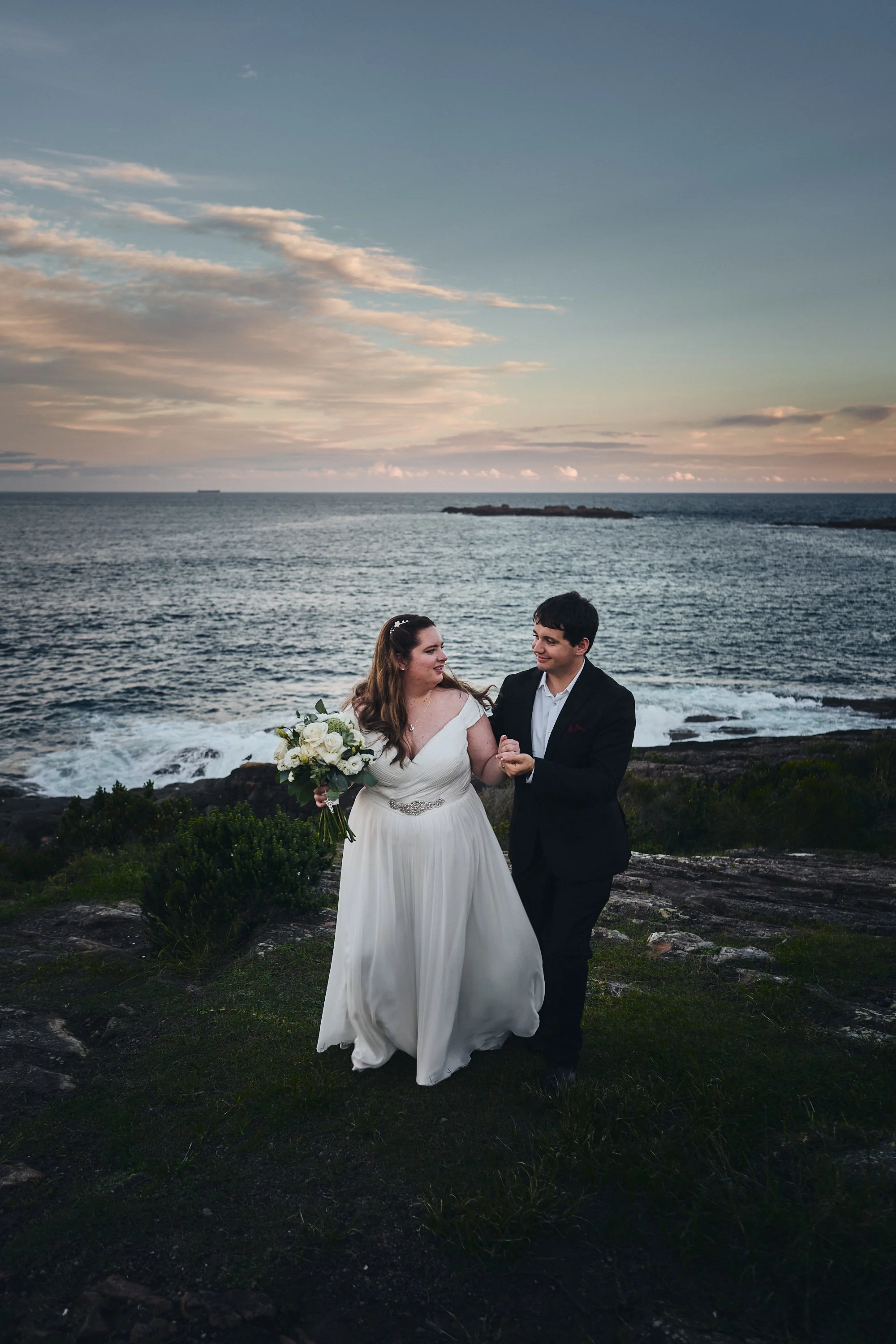 Bride and groom walking on a grassy hillside by the ocean at sunset