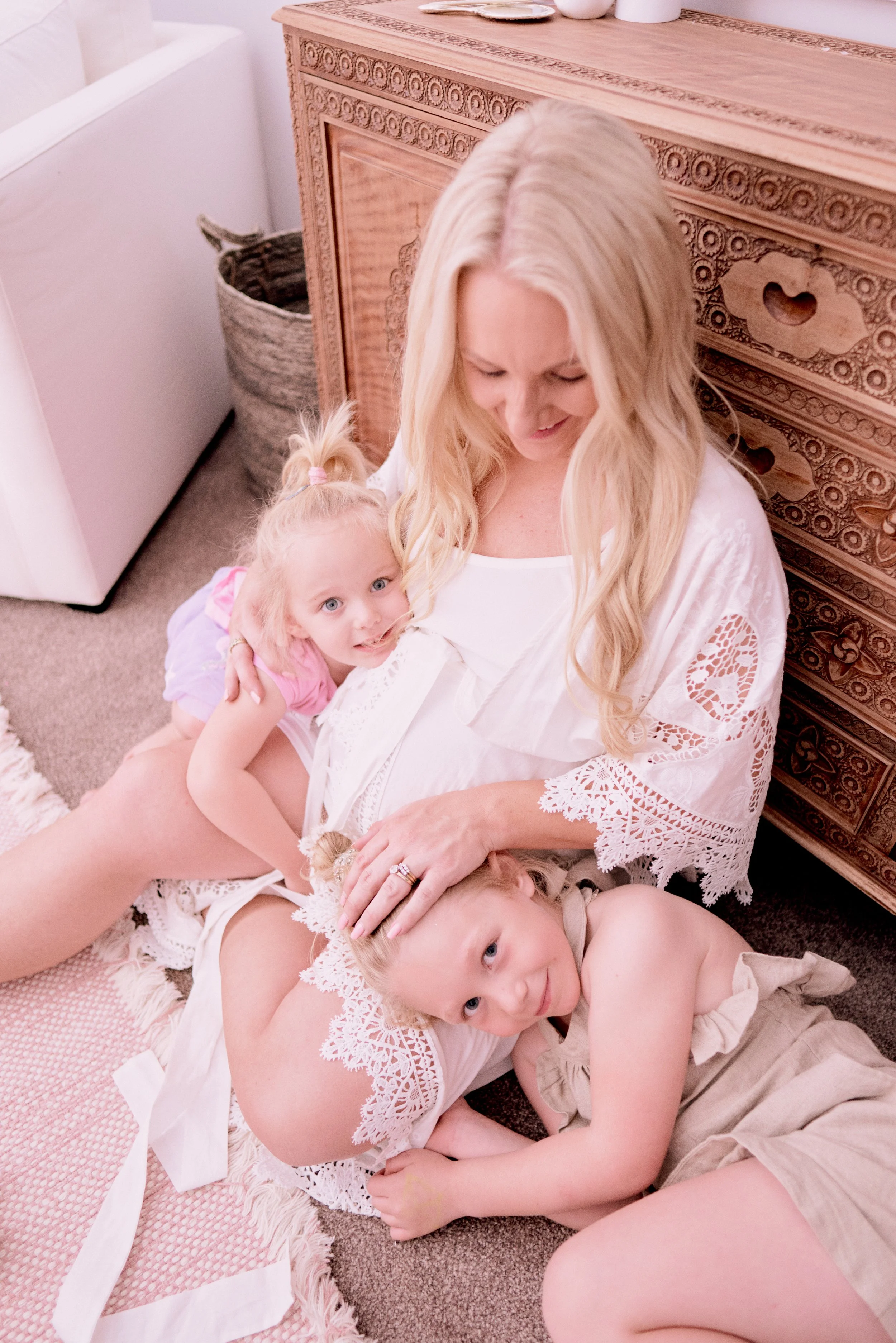 A mother sitting on the floor with two young children hugging her. They are next to a wooden dresser with intricate carvings, and the mother is wearing a white lace-trimmed robe. The children have light hair and are smiling.