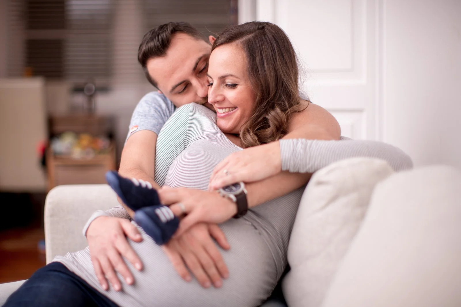Pregnant couple embracing on a couch, with small baby shoes on the woman's belly, showing happiness and anticipation.