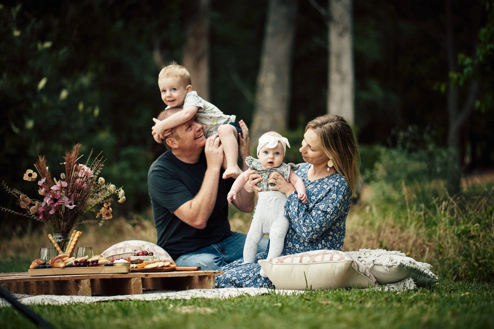 A family enjoying a picnic in a park. A man is playfully holding a young boy on his shoulders, while a woman holds a baby. A wooden pallet acts as a table, with food and drinks arranged on top. The family sits on cushions, surrounded by greenery.