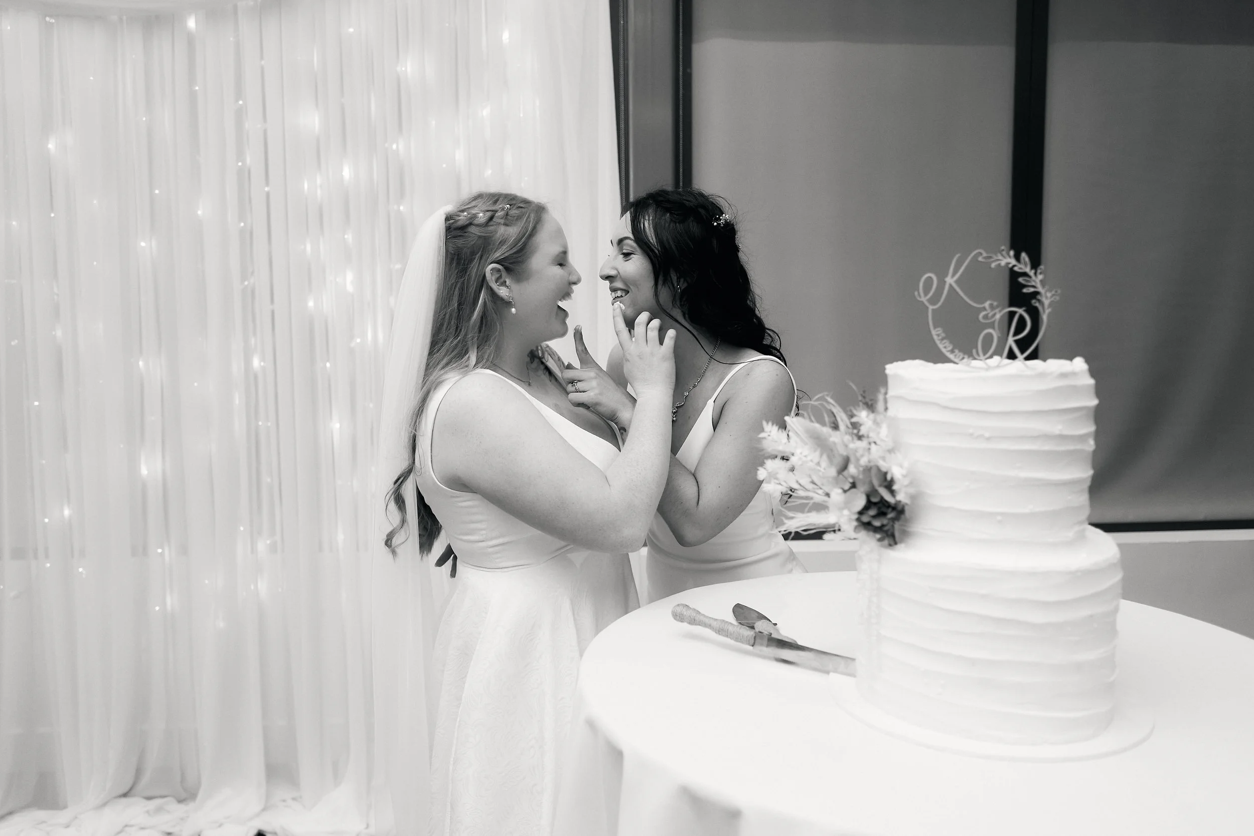 Two women in wedding dresses sharing a tender moment at their wedding reception, with a wedding cake decorated with flowers on a table nearby.