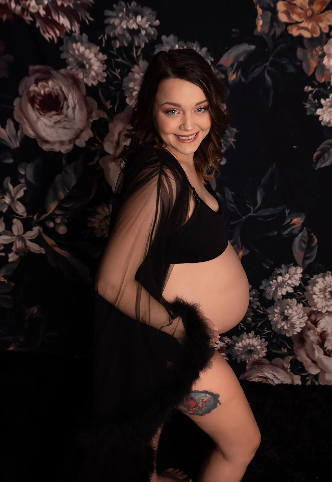 Pregnant woman smiling, wearing black outfit with sheer sleeves, sitting in front of floral black background.