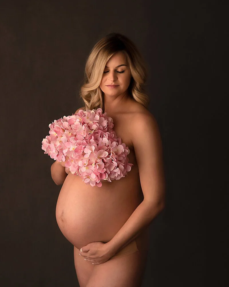 Pregnant woman with wavy blonde hair holding pink hydrangea flowers over her chest, standing against a dark background.