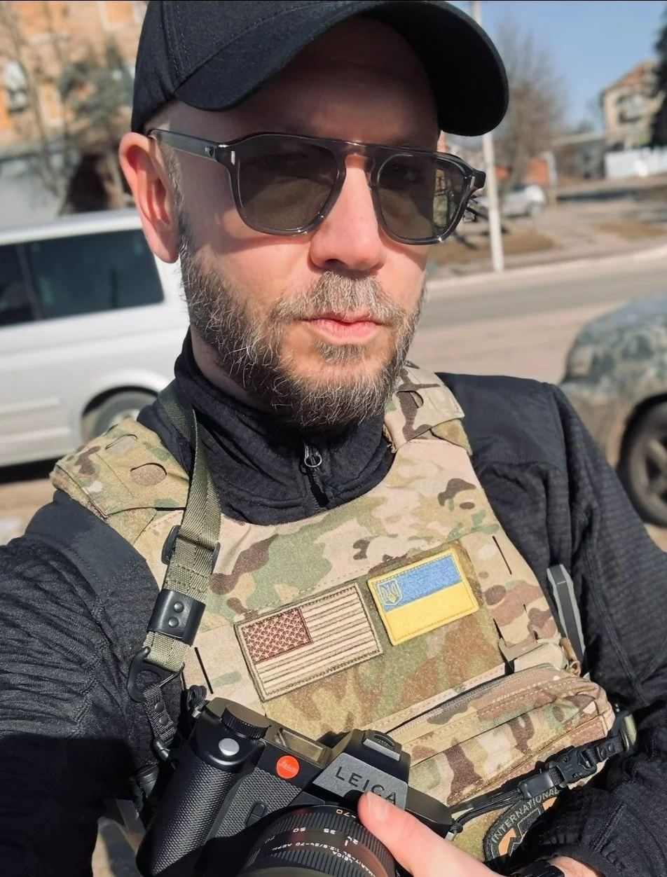 Person wearing a camouflage vest with American and Ukrainian flag patches, holding a Leica camera, wearing sunglasses and a black cap.