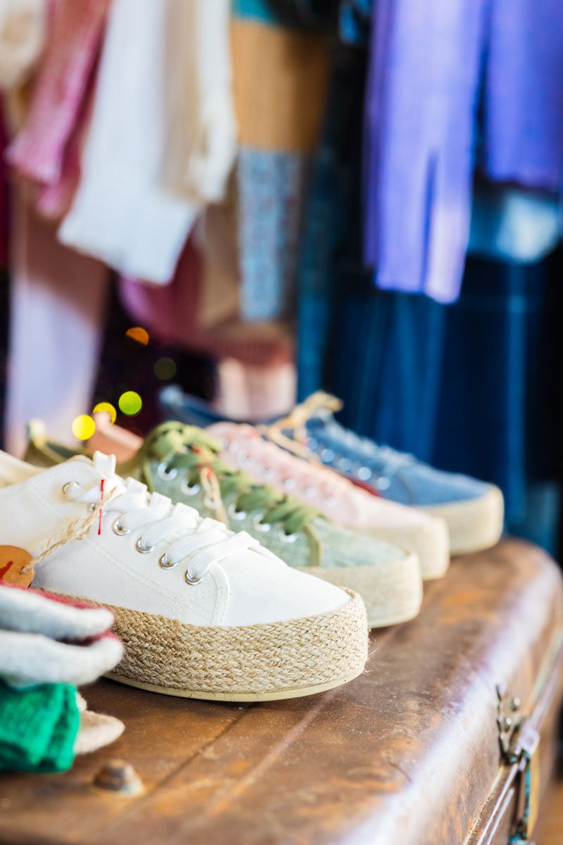 A row of shoes sitting on an old trunk with clothes hanging in the background