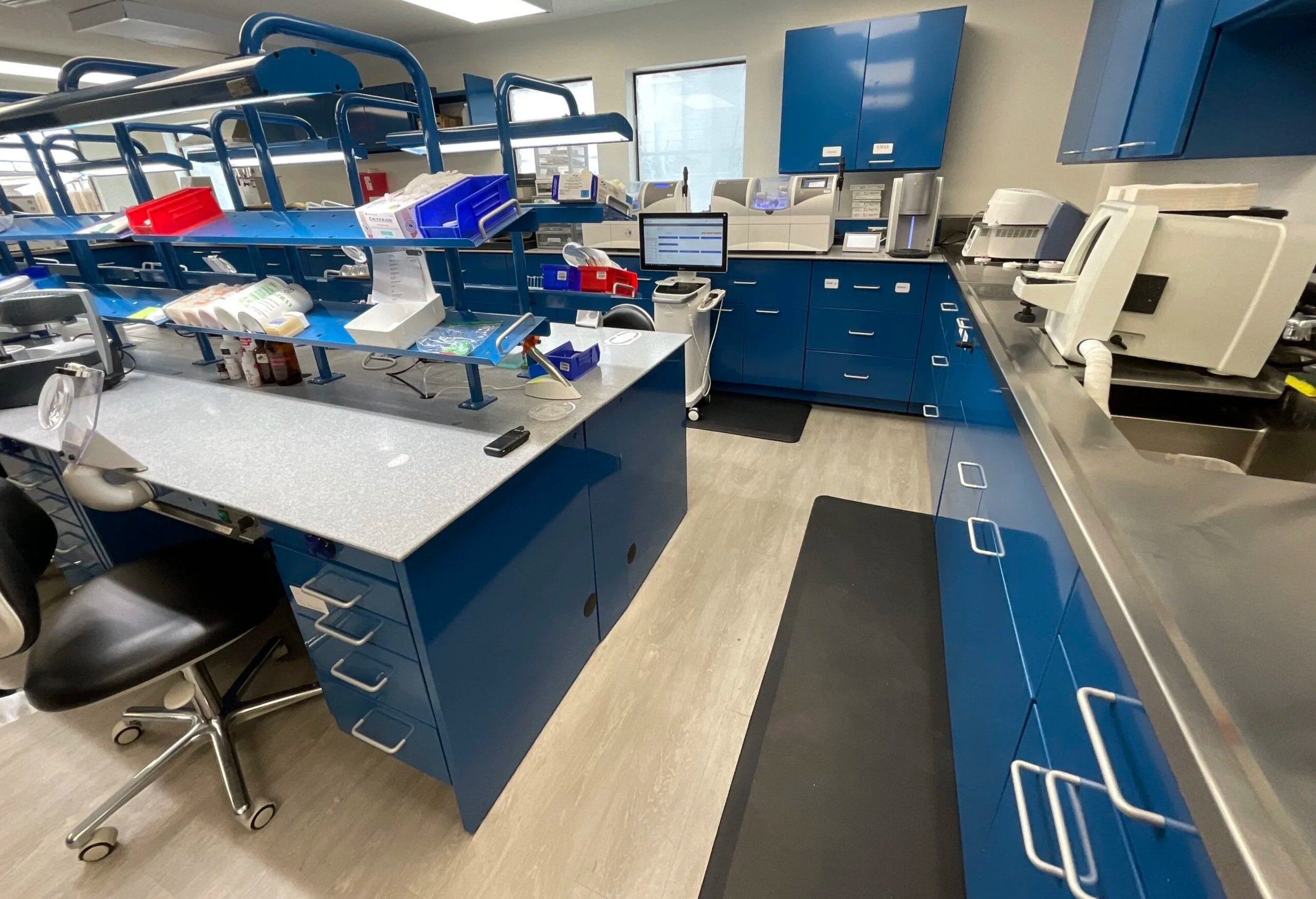 Medical laboratory with blue cabinets, testing equipment, and supplies, including a computer, lab instruments, and storage bins.