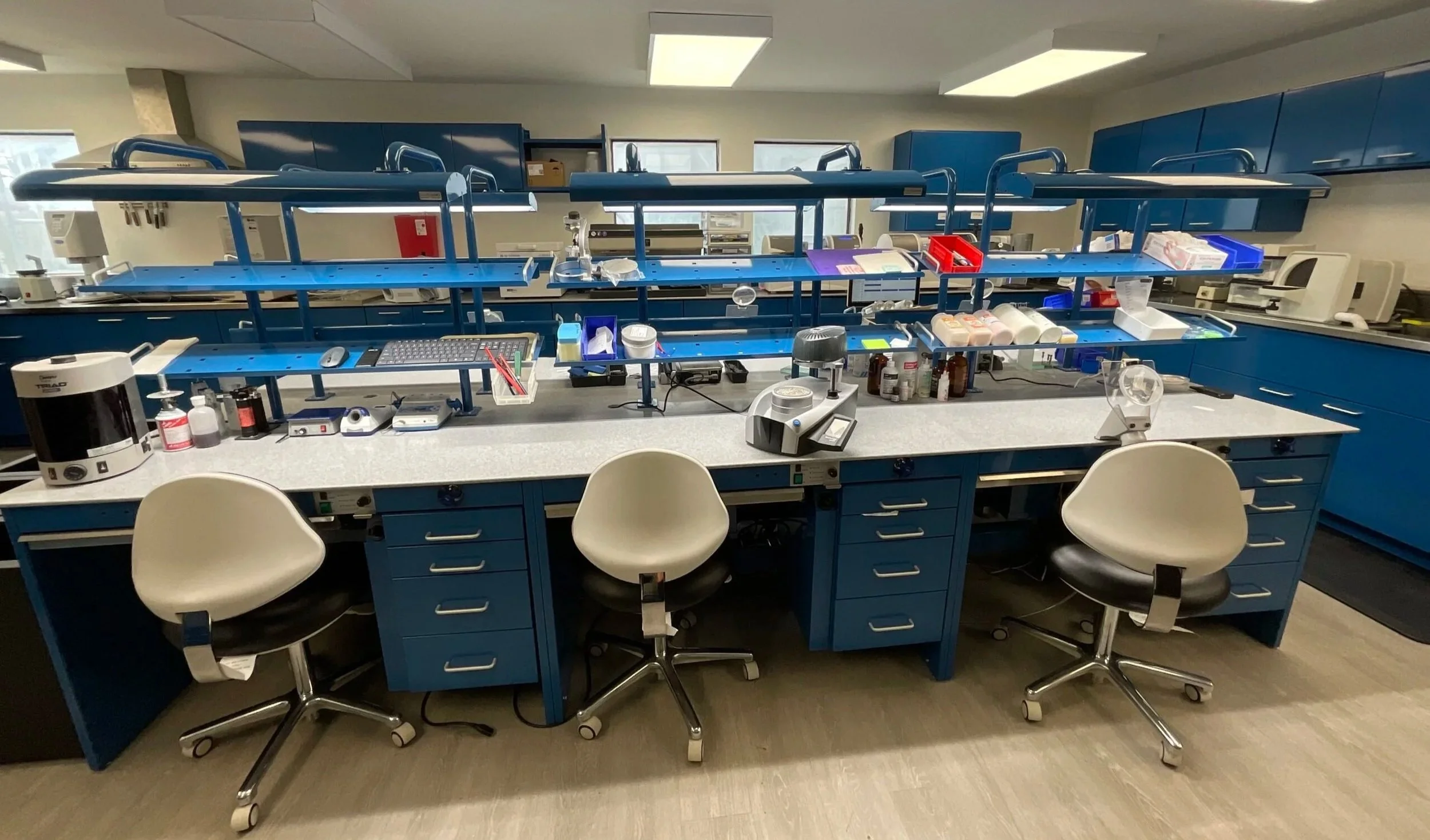 Laboratory workspace with blue cabinets, countertops, and a central blue shelving unit. Three chairs are positioned in front of the workspace, with various lab equipment, containers, and supplies on the countertops and shelves.