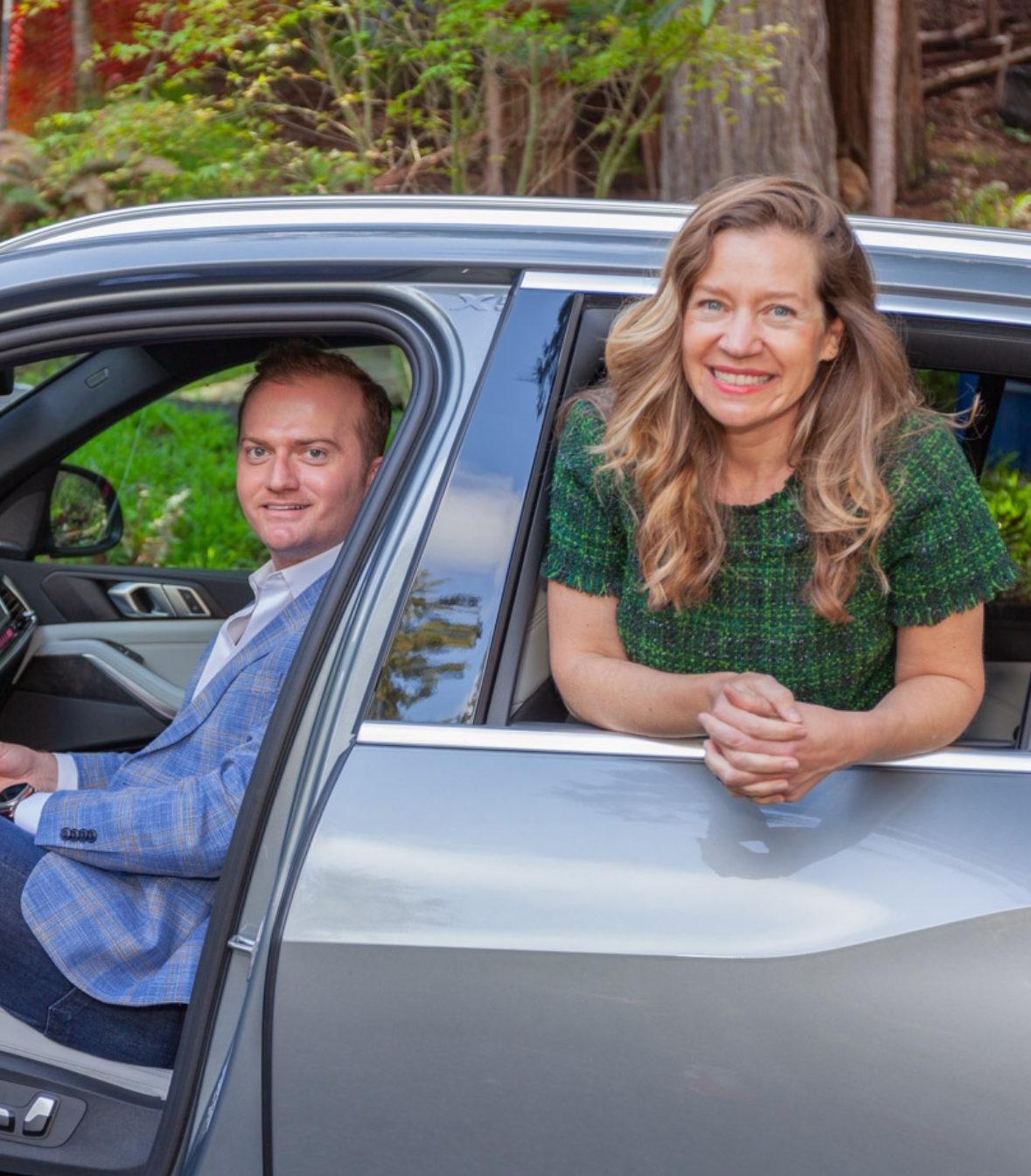 A woman with long wavy hair smiling outside a moving car with a man in the driver's seat, in a green natural setting.