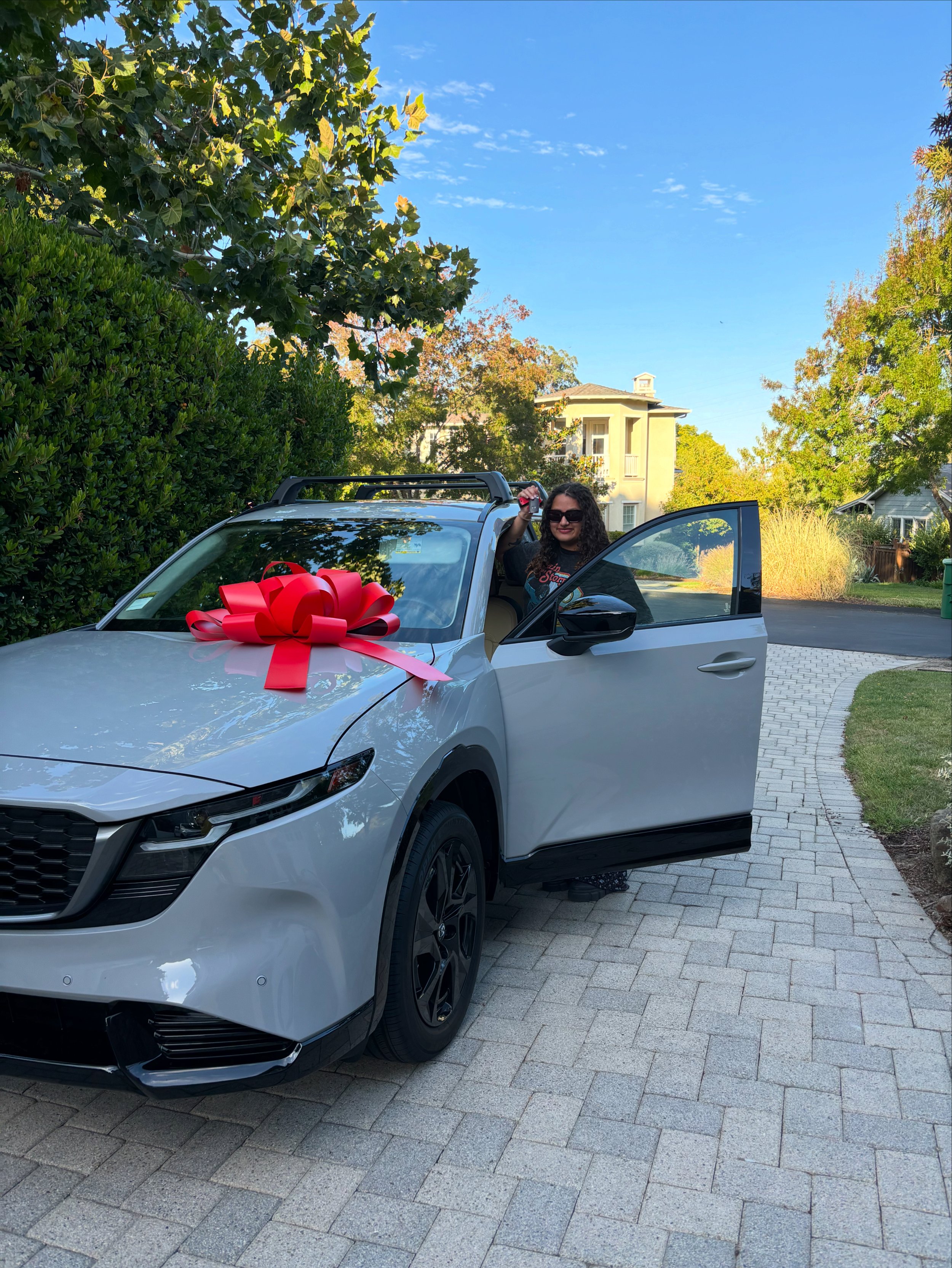 A woman with sunglasses and curly hair smiling inside a gray SUV decorated with a large red bow, parked on a paved driveway in a residential neighborhood with green trees and houses in the background, during daytime.